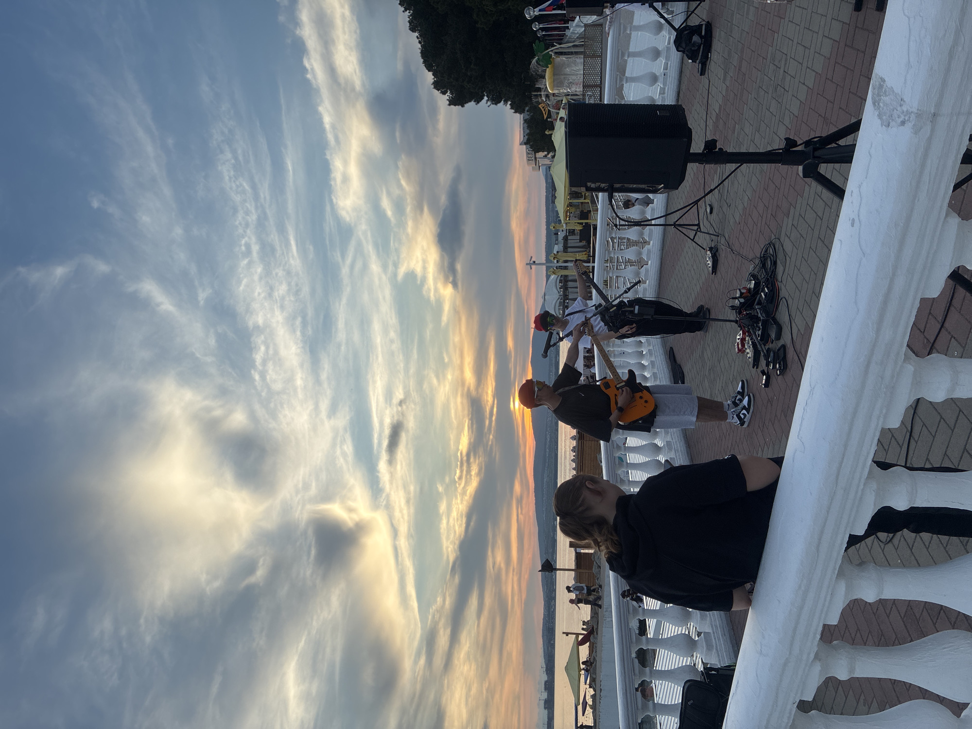 An outdoor scene at dusk features a group of people performing music near a body of water, likely a sea or large lake, with a scenic sky filled with clouds. In the foreground, a person with their back to the camera, wearing a black hooded sweatshirt and dark pants, leans on a white ornate railing. To their right, two individuals are actively engaged in a musical performance. One, wearing an orange beanie, sunglasses, a dark t-shirt, and white shorts, is playing an orange electric guitar. The other, to their right and slightly further back, is also playing a guitar. Various musical equipment, including pedals and amplifiers, are visible on the paved ground. Beyond the performers, a body of water stretches to the horizon, with a distant shoreline featuring buildings and green hills under a vibrant, cloud-streaked sky transitioning from yellow and orange near the horizon to blue overhead. The overall atmosphere suggests a public gathering or performance space by the water during sunset.