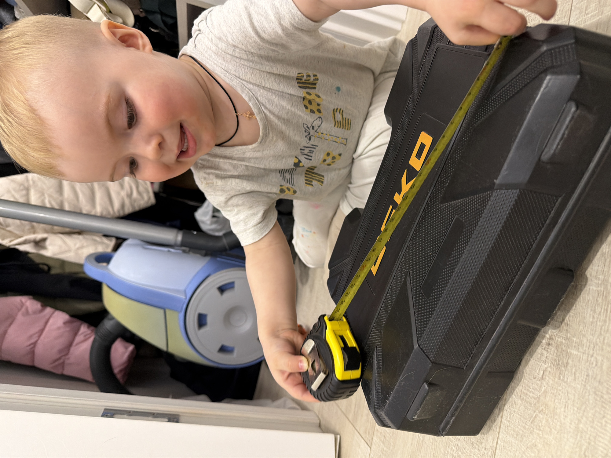 A blonde toddler in a gray t-shirt and white pants measures a black tool case with a yellow tape measure on a wood floor. A blue and yellow vacuum cleaner, clothing, and shelves are visible in the background, suggesting a closet setting. The scene does not contain enough information to determine if the location is in the city of Votkinsk, Russia.