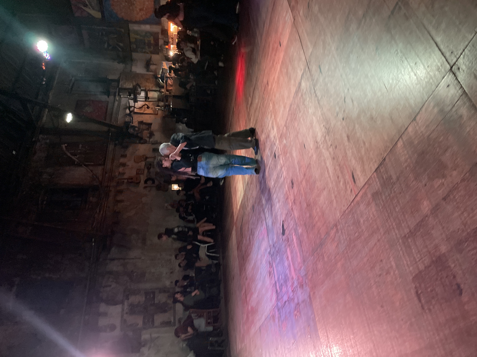 A medium shot captures a dimly lit scene with a large wooden dance floor occupying most of the frame. Two figures are positioned centrally, engaged in what appears to be a dance or embrace. One figure has gray hair and is wearing a dark jacket and khaki pants, while the other wears a black top and blue jeans. Their interaction is intimate.
Along the left side of the frame, a crowded area with people sitting at tables and standing can be observed. The background shows the interior of a building with brick walls and various decorations and lighting fixtures. The lighting is dim, with spotlights illuminating the scene and casting shadows.
The image is rotated slightly clockwise. This scene may take place in a tango hall in Buenos Aires, Argentina.
