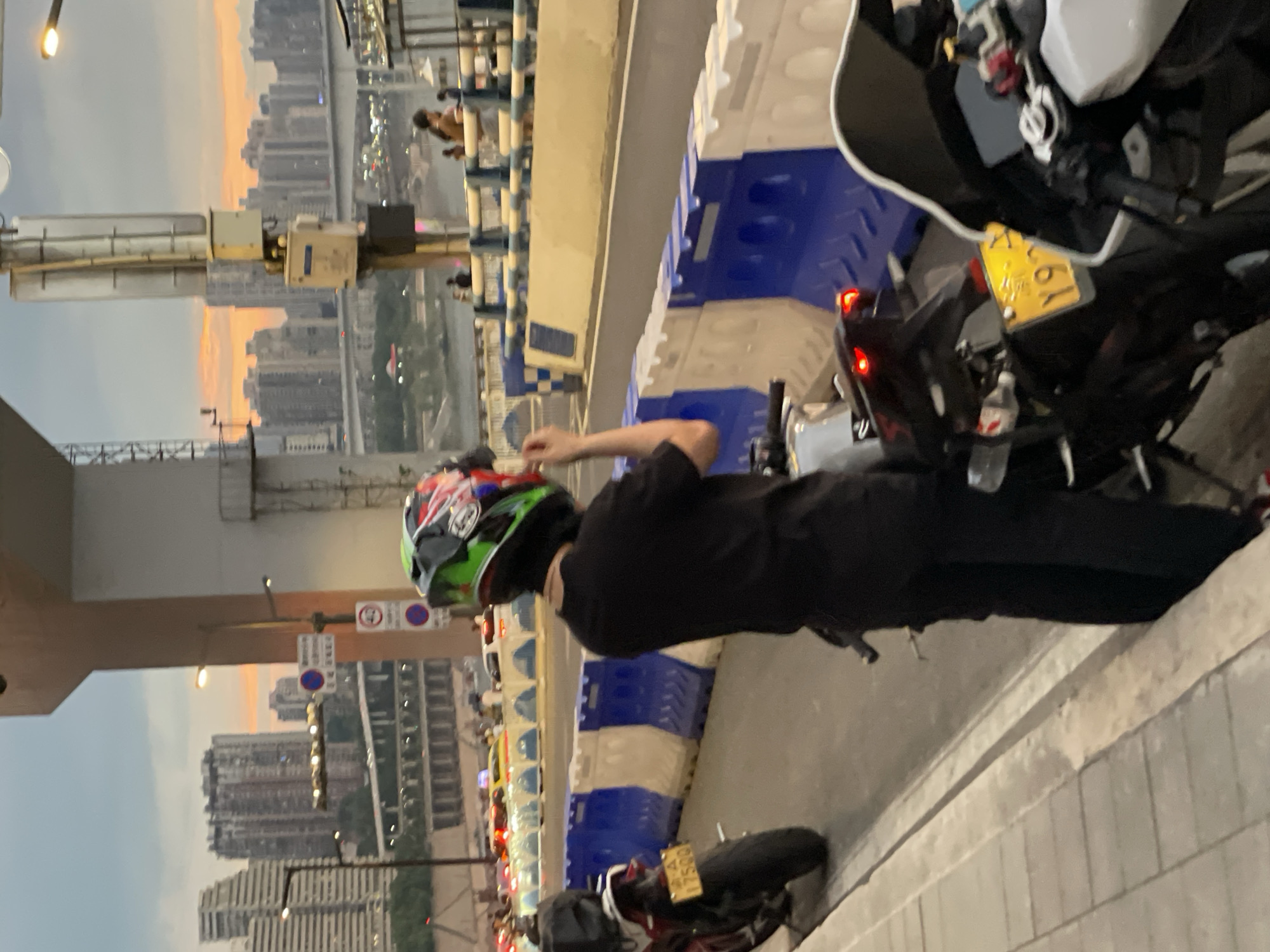 The image shows a street scene at dusk in the Yuzhong District of China. A person wearing a black t-shirt and black pants, and a colorful motorcycle helmet, stands facing away from the camera. The person appears to be adjusting their helmet. In the foreground, two motorcycles are parked on the side of the road. The motorcycle on the left is red and has a yellow license plate. The motorcycle on the right is white and has a yellow license plate with characters that are partially obscured. The background shows a cityscape with tall buildings and a bridge. Traffic is visible on the bridge and in the distance. Blue and white barriers line the street. The sky is cloudy with hints of orange from the setting sun.