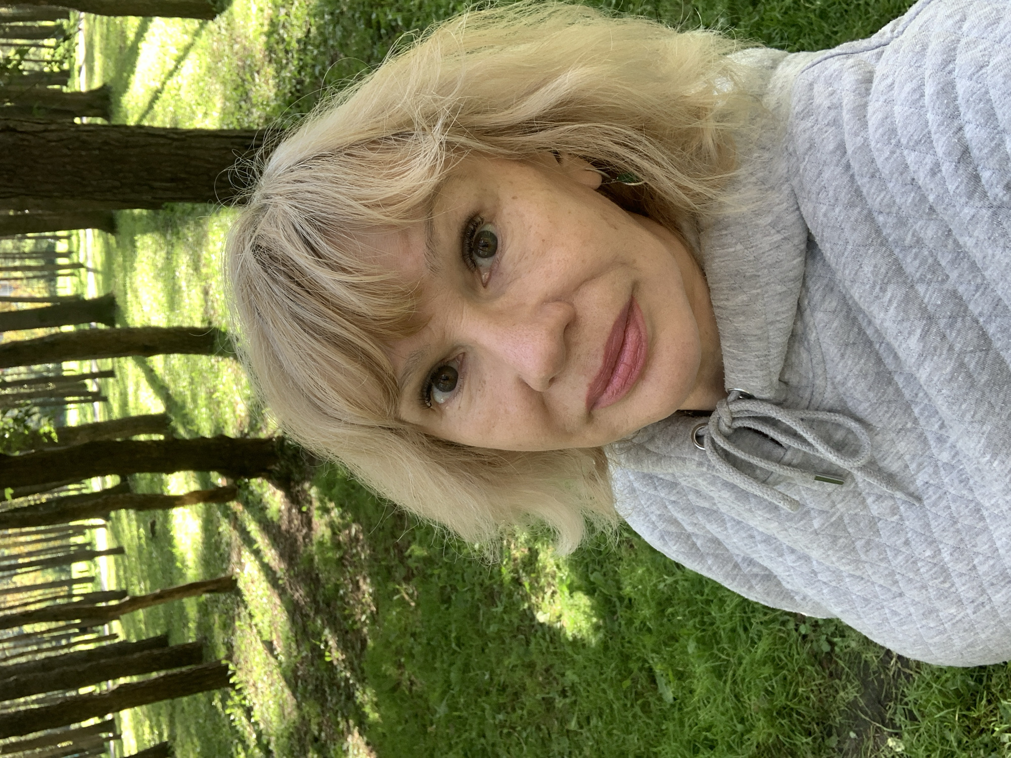 A medium shot selfie captures a woman with blonde, shoulder-length hair and light freckles on her cheeks. She is wearing a light gray quilted hoodie with a drawstring. Her eyes are brown, and her lips are subtly colored. She is positioned in a natural setting, likely a park or wooded area, with tall trees and lush green grass visible in the background, dappled with sunlight and shadows. The environment suggests an outdoor activity, possibly a walk or relaxation in nature. The image does not provide any specific information or visual cues that would indicate a location in Prokopyevsk, Russia.