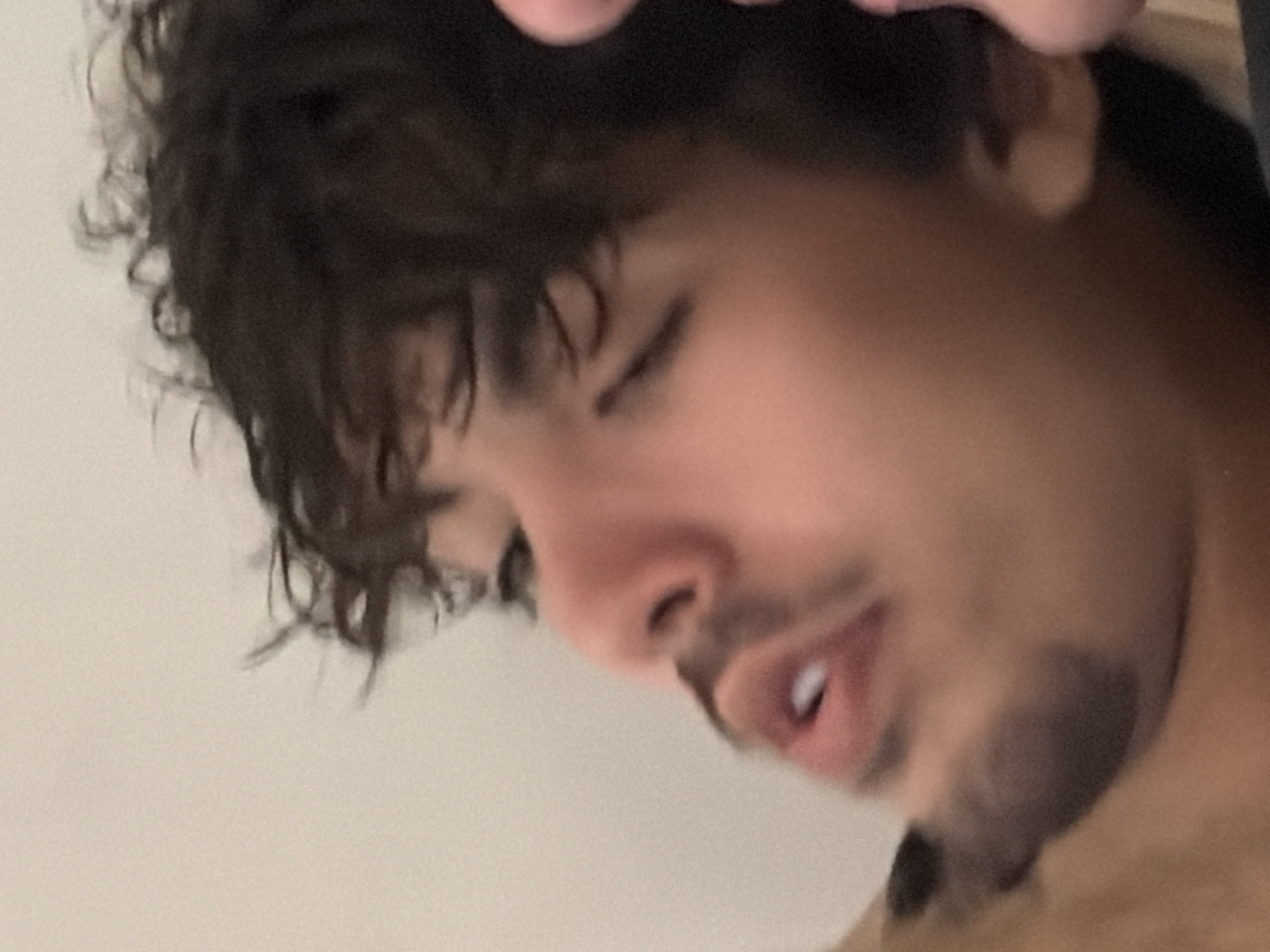 The image is a close-up, side profile shot of a young person's face. The person has dark, curly hair, dark eyebrows, and is looking downwards with their mouth slightly open, revealing a white tooth. They have a mustache and a goatee. The background is a plain, light-colored wall. The image is blurred and grainy, making it difficult to discern finer details. There is no visible indication of the person's location within Germasogeia Municipality, Cyprus, or any other specific setting.