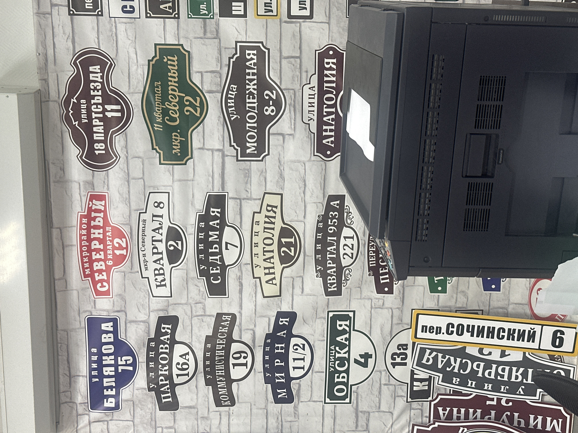 The image depicts a wall adorned with various street signs, likely for decorative or informational purposes, with a printer in the foreground. The signs are primarily in Russian and indicate street names and building numbers. Notable street names include "БЕЛЯКОВА", "ПАРКОВАЯ", "КОММУНИСТИЧЕСКАЯ", "МИРНАЯ", "ОБСКАЯ", "КВАРТАЛ 953 А", "СЕДЬМАЯ", "АНАТОЛИЯ", "18 ПАРТСЪЕЗДА", "МОЛОДЕЖНАЯ", and "МИЧУРИНА". There are also signs for "микрорайон СЕВЕРНЫЙ" and "мкр-н Северный". The printer is a dark-colored office machine with vents and buttons visible. The background is a brick-patterned wall. The signs suggest a location in Russia, and the presence of street names like "18 Парсъезда" might point to a Soviet-era urban planning influence. Without further context or explicit labels within the image, pinpointing the exact city of Novoaltaysk, Russia, is not possible. However, the style of signage and the Russian language are consistent with such a location.