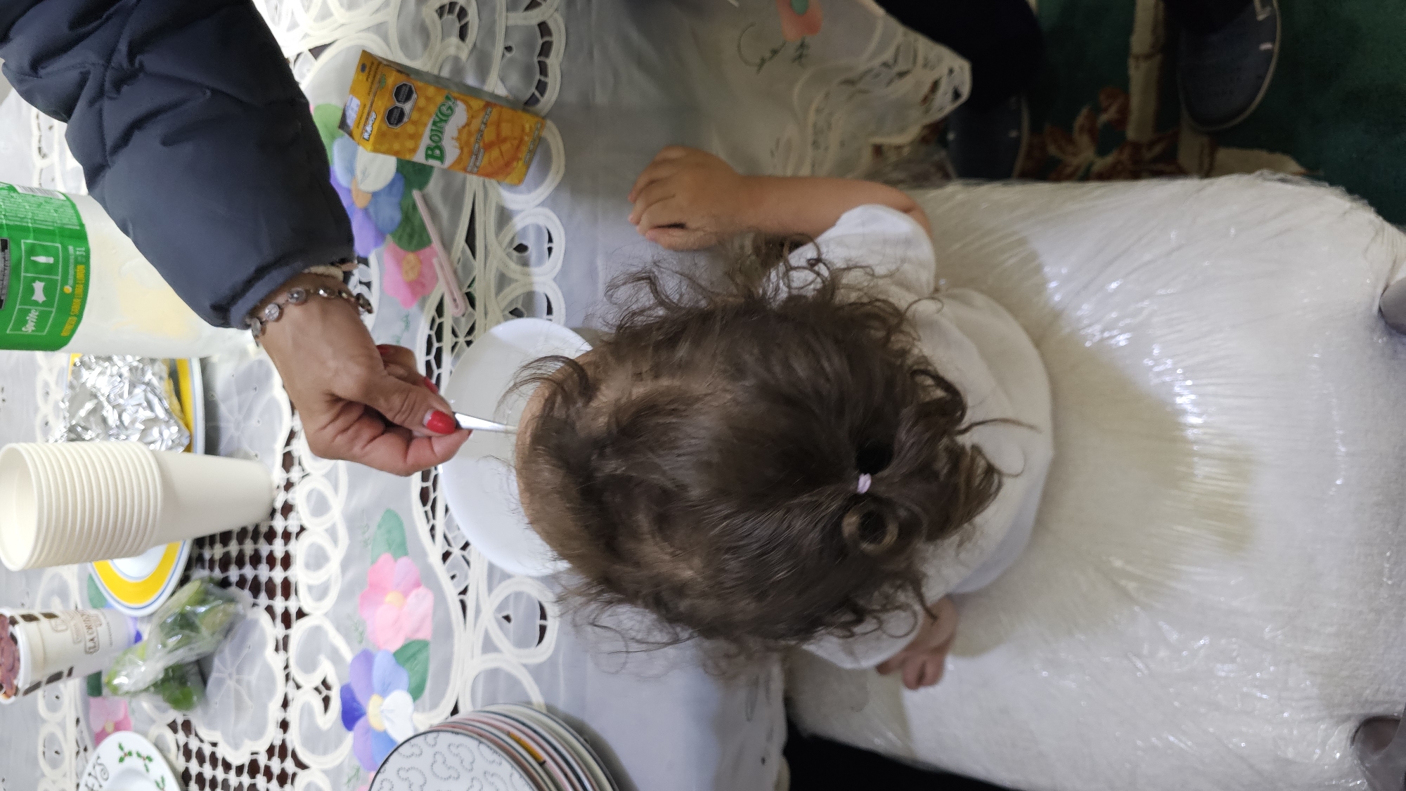The image shows an overhead view of a small child sitting at a table covered with a white tablecloth with a floral pattern. An adult hand, wearing a silver bracelet and red nail polish, holds a spoon towards the child's head, seemingly feeding them. A white bowl sits in front of the child. There are disposable cups, a bottle of Sprite, and a box of Boing! mango juice on the table. Stacked plates are also visible. The child is wearing a white shirt, and their hair is pulled back with a pink hair tie. The child is sitting on a chair that is wrapped in clear plastic. The location is San Bernardino Tlaxcalancingo, Mexico.