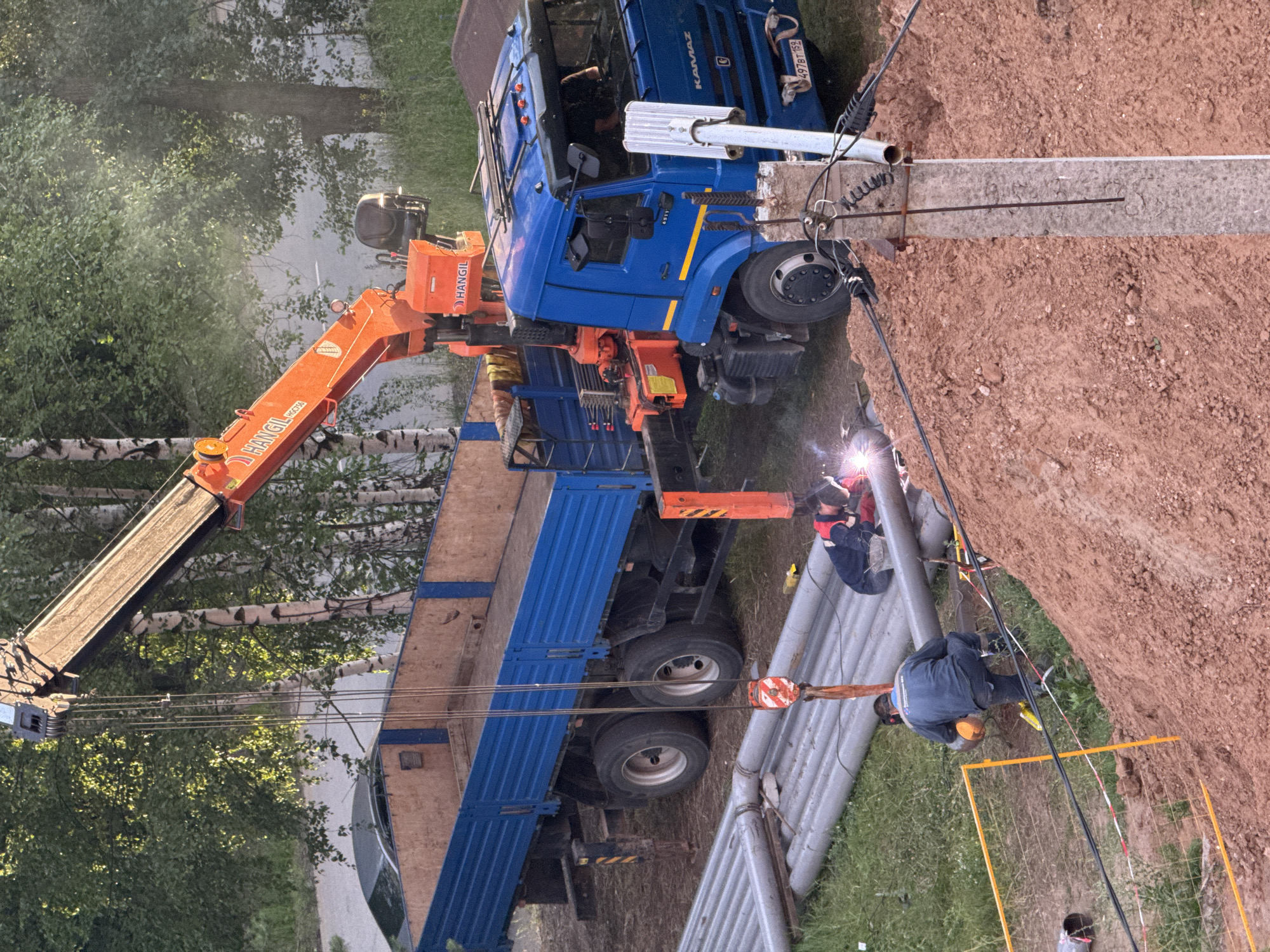 The image depicts an outdoor industrial scene in Chaikovsky, Russia, likely during daylight hours under clear weather. A blue Kamaz truck with an orange crane is parked on a dirt embankment. Two individuals are engaged in welding work on large grey pipes, one person welding in the foreground and another to the right. One welder wears a blue jacket and yellow hard hat, while the other is dressed in a red and blue work suit. The crane on the truck is extended, appearing to be involved in the pipe work or material handling. A section of the pipe appears to be a concrete pipe. A yellow safety fence is visible in the lower right corner. The surrounding environment includes trees and greenery, suggesting a partially developed or construction area. The truck has a license plate with the text "497BT 159". The crane is branded with "HANGIL".