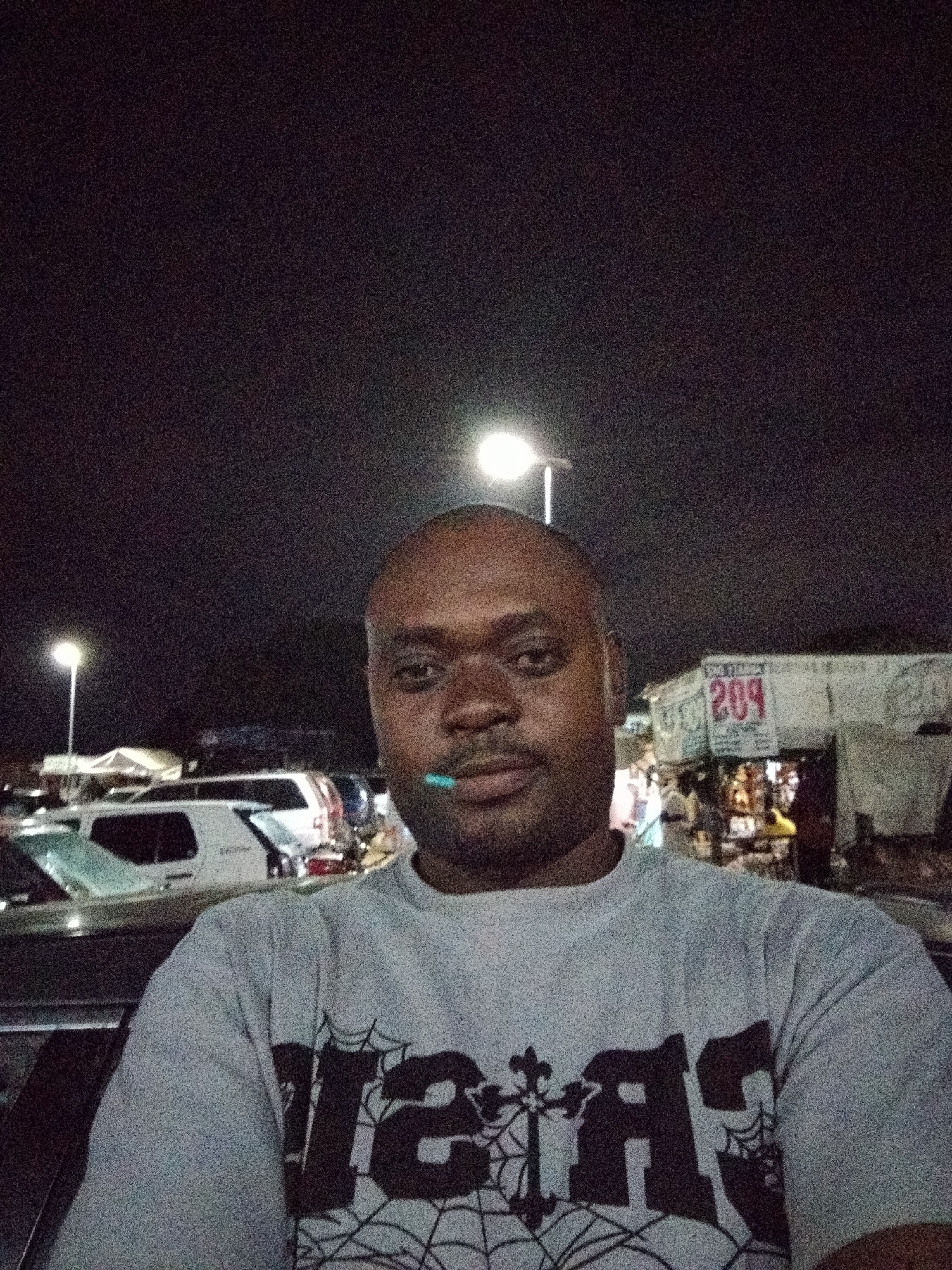 A grainy, nighttime outdoor portrait captures an adult male in the foreground, positioned in the city of Pasere, Nigeria. He has a dark complexion, a bald head, and dark stubble on his face, with a small light-blue object held between his lips. He is wearing a light grey crew-neck t-shirt adorned with a dark graphic comprising a stylized cross, spiderweb patterns, and partially visible text that appears to read 'CRAS'. The man gazes directly into the camera.
The background is dimly lit by artificial sources, including two overhead streetlights: one to the left and another higher on the right. Several vehicles are parked behind him, notably a light-colored SUV on the left. To the right, obscured commercial structures or market stalls are visible, one featuring a prominent red sign with the number '209'. The sky is dark, indicative of nighttime, and the entire scene exhibits significant image noise.