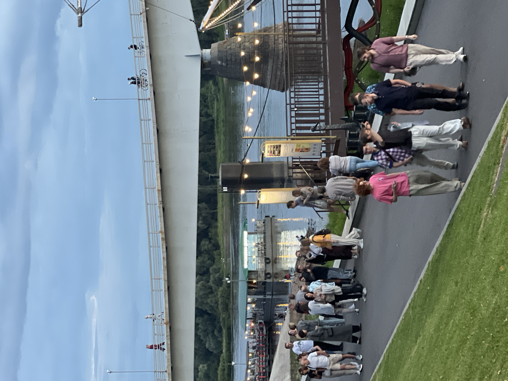 In Veliky Novgorod, Russia, an outdoor scene unfolds beneath a cloudy twilight sky, featuring a prominent bridge arching over a wide river. A crowd of people gathers on a paved walkway alongside the riverbank.
On the bridge's walkway, two individuals are seen walking in separate directions, and further along, two people are riding bicycles.
Below the bridge, on the river, a floating structure resembling a restaurant or cafe is illuminated by numerous lights. A pier extends from the riverbank, with a railing and decorative string lights. A second, larger boat with lights is docked to the right.
The main group of people are on the paved walkway in the foreground. They are dressed in casual attire. Some are standing and conversing, while others are walking. A prominent group of five men are walking towards the viewer. One man in a pink t-shirt is walking away from the viewer on the far right. A woman in a yellow top is also in this group.
The environment includes lush green grass along the left side of the walkway and the river with its reflections of lights on the right. The sky is a mix of gray clouds and lighter blue patches.