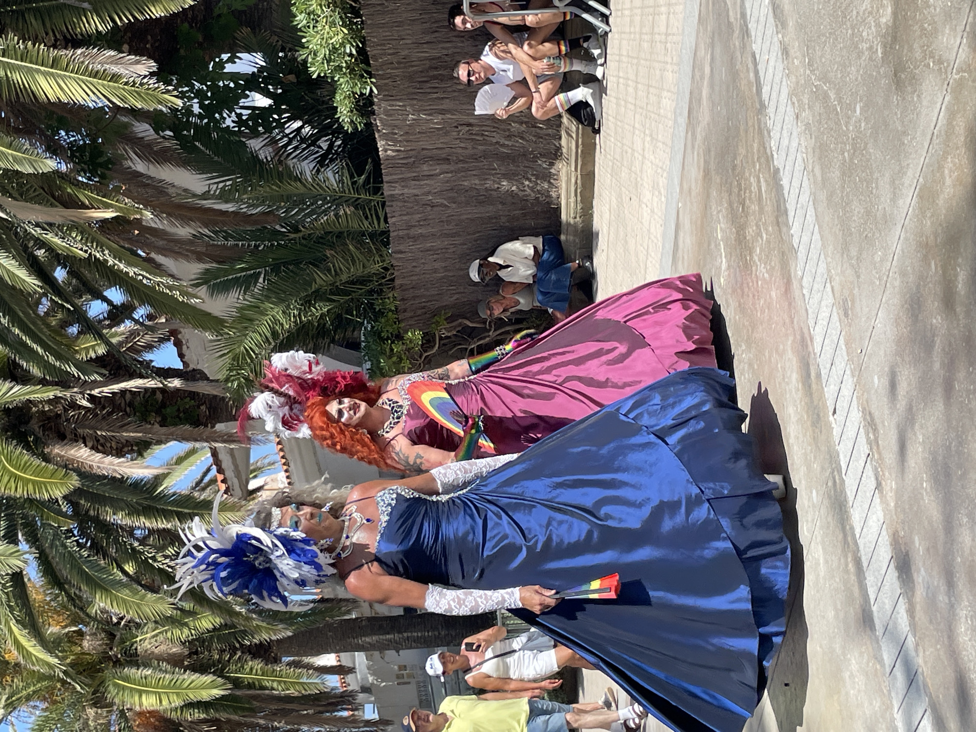 The image depicts a vibrant street scene in Sitges, Spain, likely during a Pride event. Two individuals in elaborate drag attire, adorned with large feathered headdresses and opulent gowns, are the central focus. The person on the left wears a shimmering navy blue gown and white lace gloves, while the person on the right is in a magenta gown. Both are holding small rainbow-colored fans.
In the background, palm trees line the street, casting shadows on the paved ground. Several spectators are seated along a low wall, some fanning themselves, while others stand and observe the procession. One man in the foreground wears a yellow shirt and shorts, and a woman behind him in a white tank top and shorts holds up a mobile phone, likely capturing the event. The architecture visible suggests a Mediterranean setting. The overall atmosphere is celebratory and sunny.