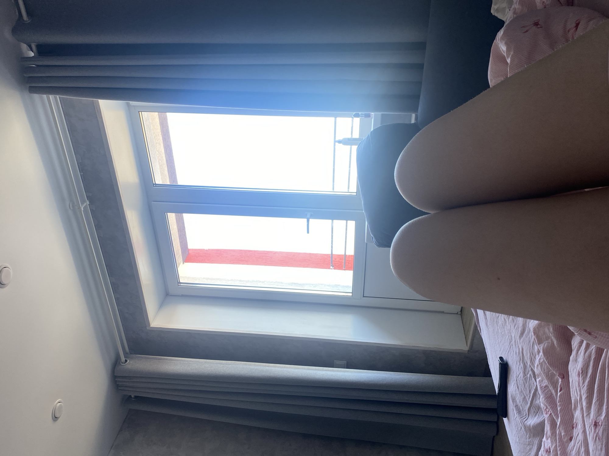 The image captures an indoor scene, likely a bedroom or living room, from a low perspective, rotated 90 degrees counter-clockwise. In the foreground, a person's bare legs, specifically the thighs and knees, are visible, resting on pink and white patterned bedding. The legs extend from the right side of the frame.
A large, modern window with white frames dominates the left and central portion of the background. The view outside the window is extremely bright, indicating strong daylight. Below the window, gray pleated Roman blinds are visible, partially drawn. Similar gray curtains are present above the window, also partially covering the top part of the frame. A dark piece of furniture, possibly a sofa armrest or headboard, is partially seen behind the legs, near the right side of the window. On the white wall to the far left, two recessed circular light fixtures are visible. Below the window, a windowsill and a darker, textured wall or floor material are present. A small black rectangular object, likely a phone or remote control, rests on the pink bedding near the bottom right.
The activity depicted is a person resting or relaxing indoors during the daytime in Zubovo, Russia. No visible text is present.