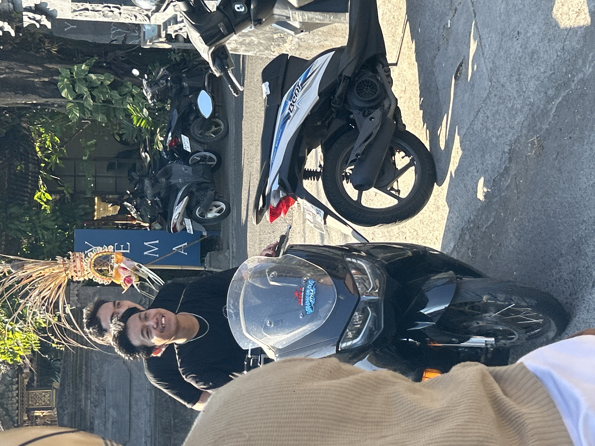 In North Kuta, Indonesia, two young men are visible. The man in the foreground, wearing a black t-shirt and a silver chain, is smiling broadly and sitting on a black scooter. Behind him, another young man, also wearing a black t-shirt, is partially obscured. A decorated straw ornament is positioned between the two men, and a blue sign with white lettering that appears to read "YEMA" is visible in the background. Several other scooters are parked on the paved street, with lush green foliage and trees surrounding the area, suggesting a tropical urban environment. The lighting indicates it is daytime, with sunlight casting shadows on the ground.