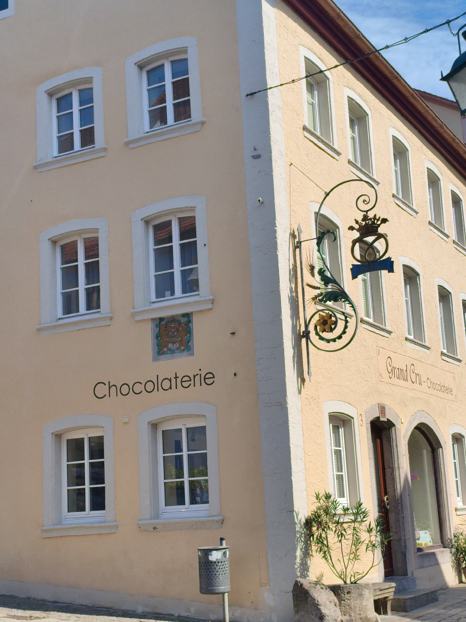 This is a medium-shot, eye-level, outdoor photograph of a building in Rothenburg ob der Tauber, Germany. The building, painted a pale yellow, has several windows with white frames. A sign reading "CHOCOLATERIE" is mounted on the wall between two windows on the lower floor. Above this sign, a decorative wrought-iron element with a crown, a ribbon, and a flower is suspended from the building. Further down the street, a sign reads "Grand Cru - Chocolaterie". To the right of the entrance, a small tree grows in a stone planter. A silver trash can with a perforated design is visible near the corner of the building. The sky above is blue with some white clouds.