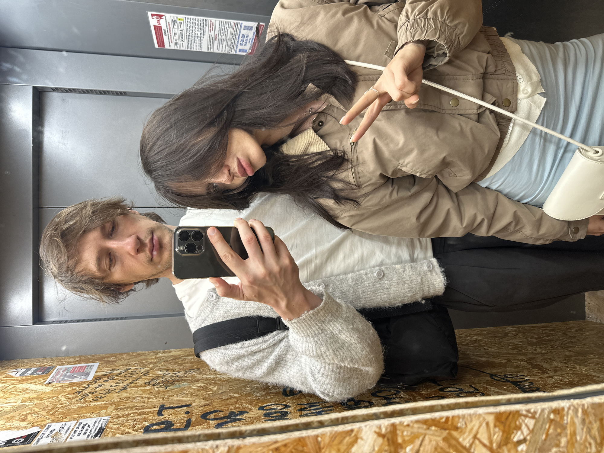 In an elevator, a man and a woman take a mirror selfie. The man is on the left, wearing a light gray cardigan over a white t-shirt and black pants. He holds a smartphone in his right hand, angled to capture the reflection. The woman is on the right, wearing a tan jacket over a light blue skirt and a white top. She is making a peace sign with her right hand, and her hair is obscuring part of her face. The elevator walls are paneled with a textured wood-like material on the left and a metallic gray on the right. There are various posters and writing on the wooden wall, and a notice is visible on the metallic wall. A black backpack is slung over the man's left shoulder, and the woman carries a white purse.