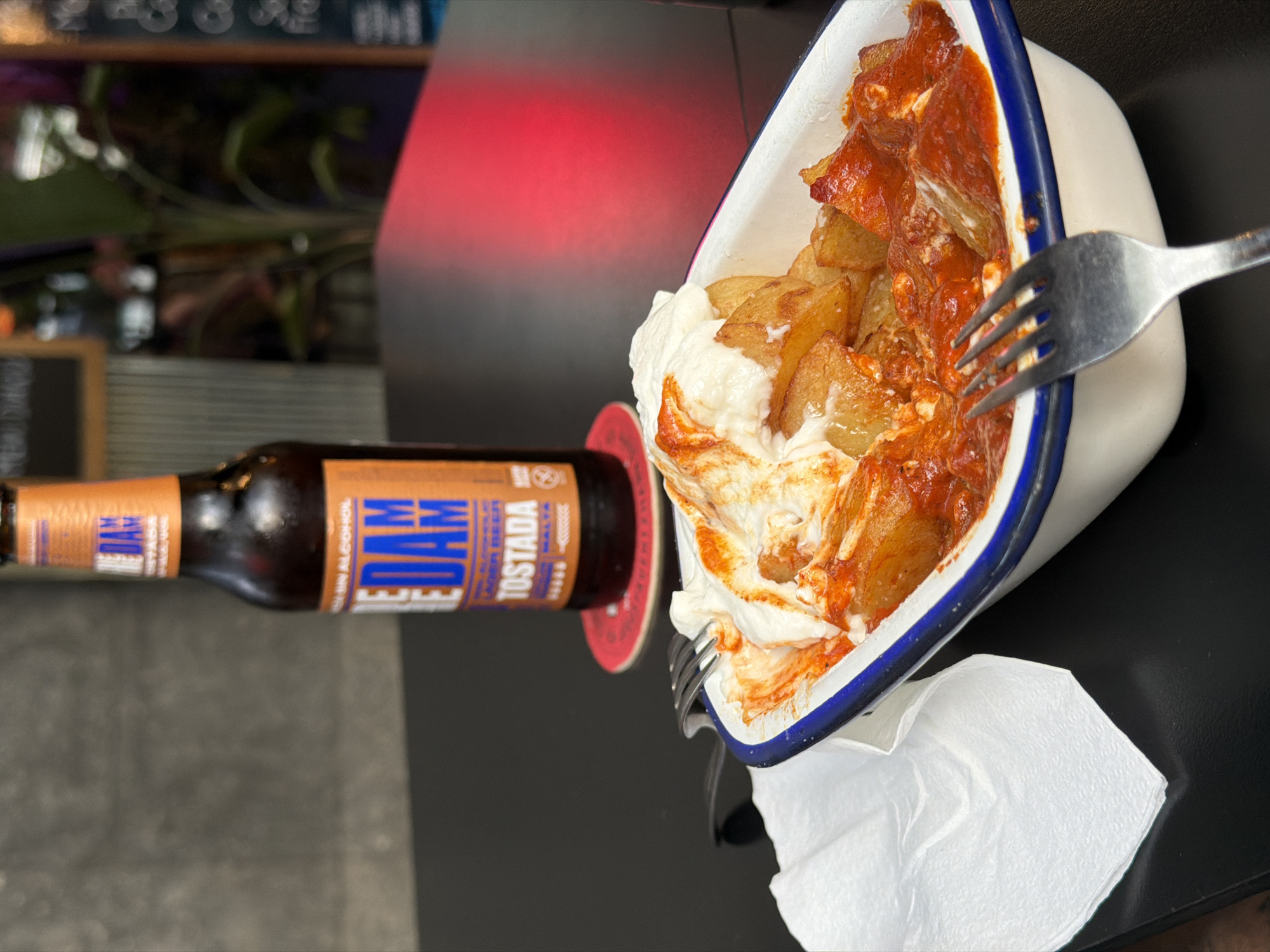 A close-up, top-down view shows a serving of patatas bravas with a bottle of beer. The patatas bravas are served in a white enamel dish with a blue rim. They are cut into wedges and coated in a red sauce and white aioli. A fork is piercing the dish and resting on the potatoes. The dish appears to be on a dark surface, possibly a table. To the left of the dish is a brown glass bottle of beer with an orange and blue label. The text on the label reads "THE DAM TOSTADA MALTA". The bottle is lying on its side, and a red coaster is visible beneath it. In the background, a red light illuminates a blurred area, suggesting an indoor setting with ambient lighting. The overall scene depicts a casual meal, likely at a bar or restaurant in Barcelona, Spain. The focus is on the food and drink, suggesting a moment of enjoyment.