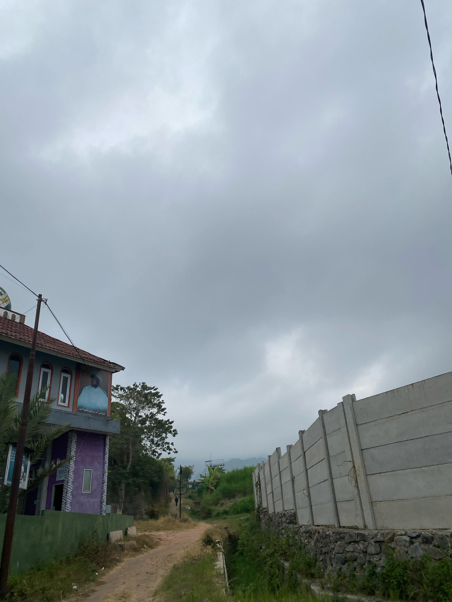 An overcast sky looms over a rural scene in Unknown, Unknown. On the left, a multi-story building with a distinctive purple lower section and a tiled roof stands beside a dirt path. A large portrait of a man in a blue robe is affixed to the upper portion of the building, and a green fence runs along its base. To the right, a long, gray concrete wall, supported by angled beams and a stone foundation, stretches into the distance. Vegetation, including grass and trees, grows alongside the path and the wall. In the background, distant green hills are visible under the cloudy sky. A single person is walking on the dirt path in the middle ground.