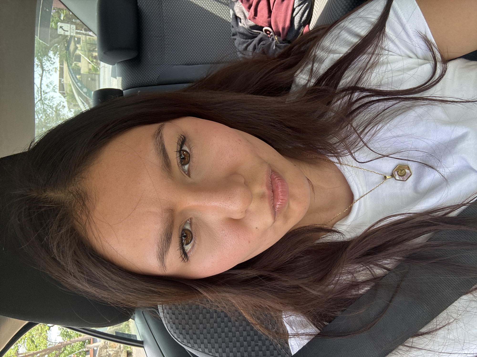 A woman with brown hair and eyes is in a car, secured by a black seatbelt and wearing a white shirt and a gold necklace. The car's interior is grey. Outside the car window, there are trees, a building, and a street sign, suggesting the photograph was taken in Oaxaca City, Mexico.