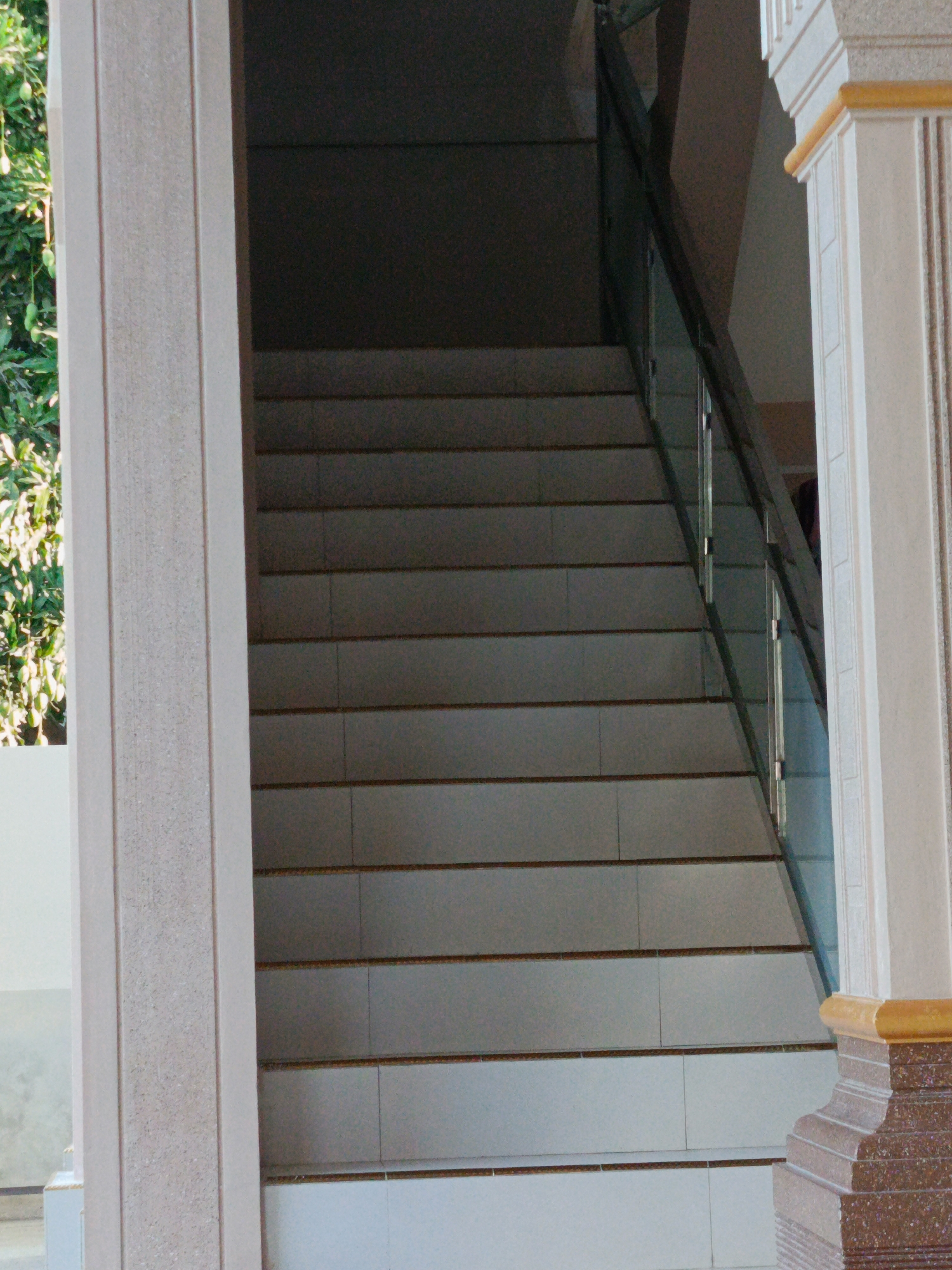 The image shows a set of outdoor stairs with a glass railing. The stairs appear to be made of stone or tile and are illuminated by natural light. A large decorative pillar is visible on the right side of the frame. The background shows lush green foliage, suggesting a tropical environment. The scene is located in Kuta, Indonesia, likely in a public area or near a building.