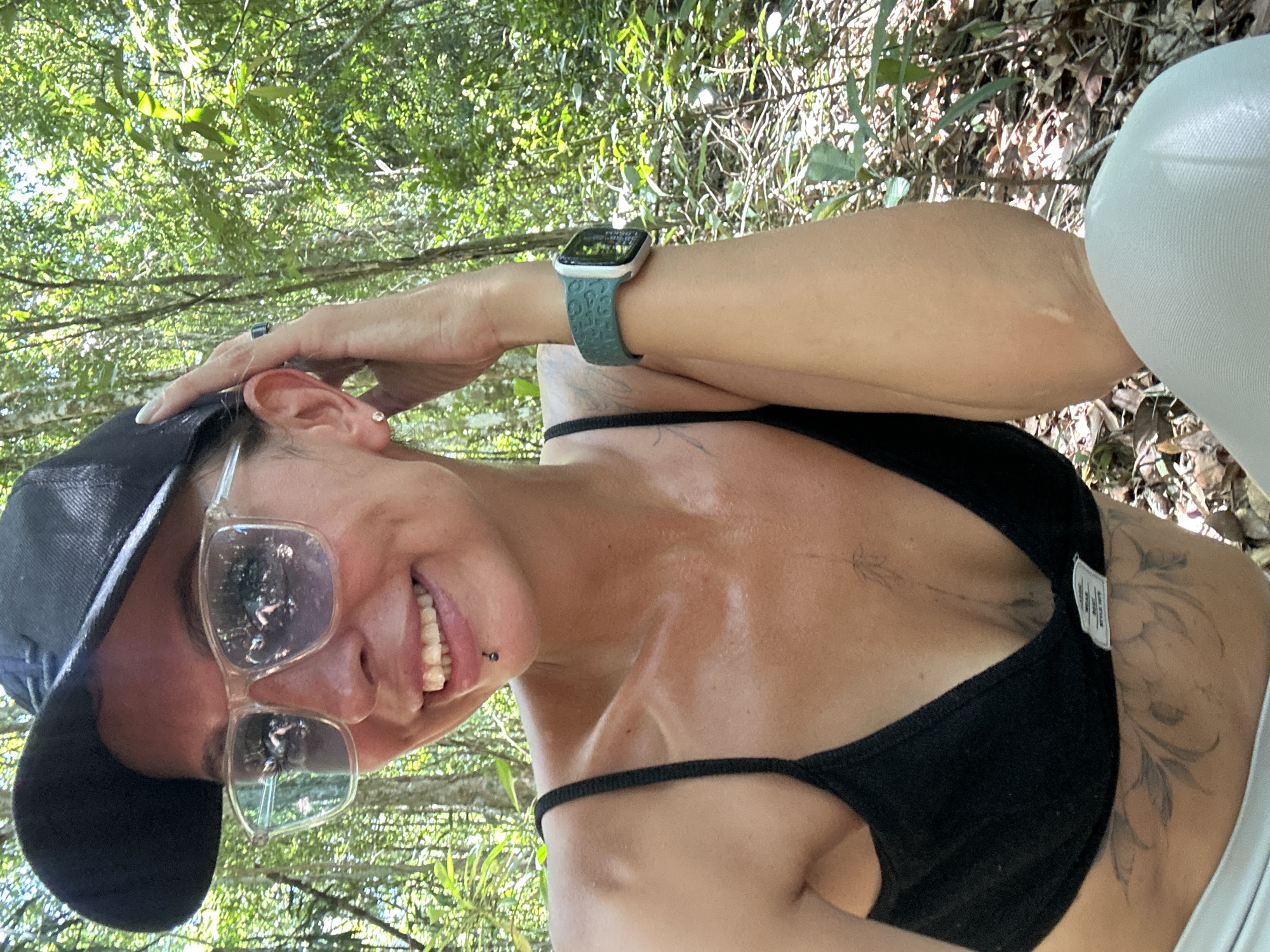 In a lush green jungle setting, a woman with sunglasses and a black baseball cap is captured in a close-up, outdoor shot. Her left hand is raised, touching the side of her head, while her right arm rests on her leg, showcasing a smart watch with a teal leopard-print band. She is wearing a black sports bra and light-colored leggings. The background is filled with dense foliage and trees, suggesting a tropical environment. The woman has a lip piercing and a few visible tattoos on her torso. Sunlight filters through the leaves, creating a dappled effect. There is no indication of the city of Ko Samui District, Thailand, in the provided image.