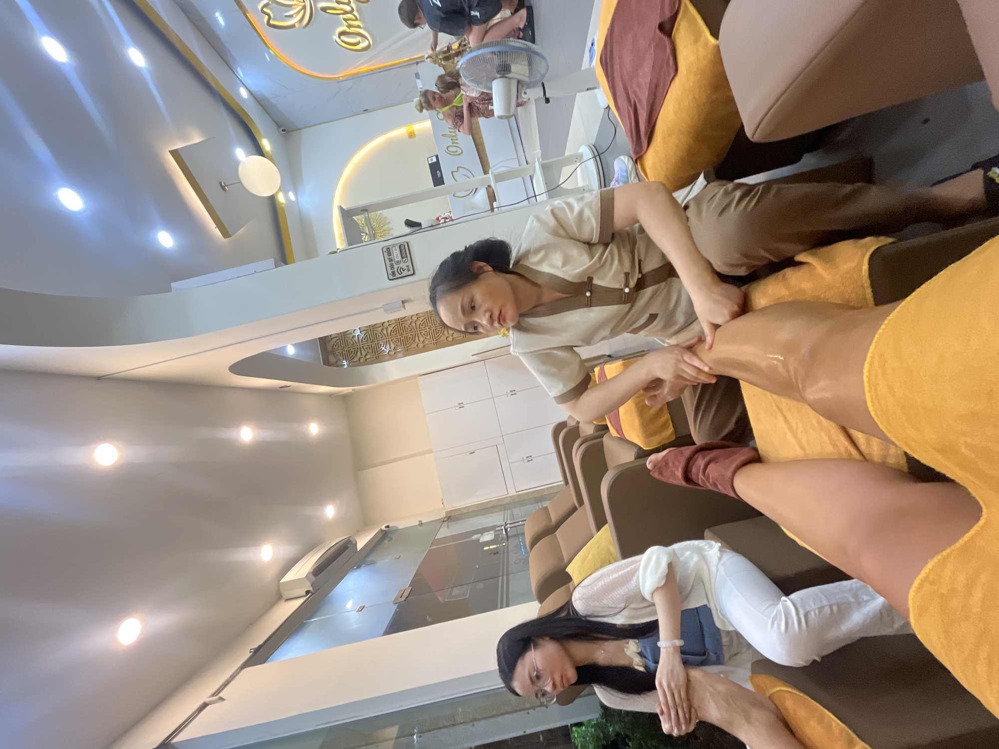 This media file depicts an indoor scene, likely a spa or massage parlor in Nha Trang Ward, Vietnam. The dominant subjects are several individuals, primarily women, receiving or providing massage services. One woman, dressed in a light brown uniform, is actively massaging the leg of another person who is lying on a massage couch, covered with a bright yellow fabric. The client receiving the massage is also partially visible, with her leg extended and resting on a cushion.
In the background, a row of similar massage couches or chairs is visible, suggesting multiple clients are being attended to. Another woman, wearing glasses and a white cardigan over a blue garment, is seated on one of these couches, with her hands clasped in front of her. Further in the background, a fan is positioned near a person who appears to be giving a massage, and there are other individuals in the distance, some receiving services.
The setting is illuminated by bright ceiling lights, giving a clean and well-lit ambiance. The walls are painted in light colors, and there are decorative elements like a golden logo with flowing script, and patterned wall accents. Some white cabinetry with doors is also visible on the wall. The overall mood seems relaxed and professional, indicative of a spa environment.
There is visible text in the image, including the golden script logo which appears to be "Only U", and some Vietnamese text on a sign that reads "KHÔNG CHẠM QUÝ KHÁCH". The bright lighting and indoor setting suggest the time of day is irrelevant to the activity.