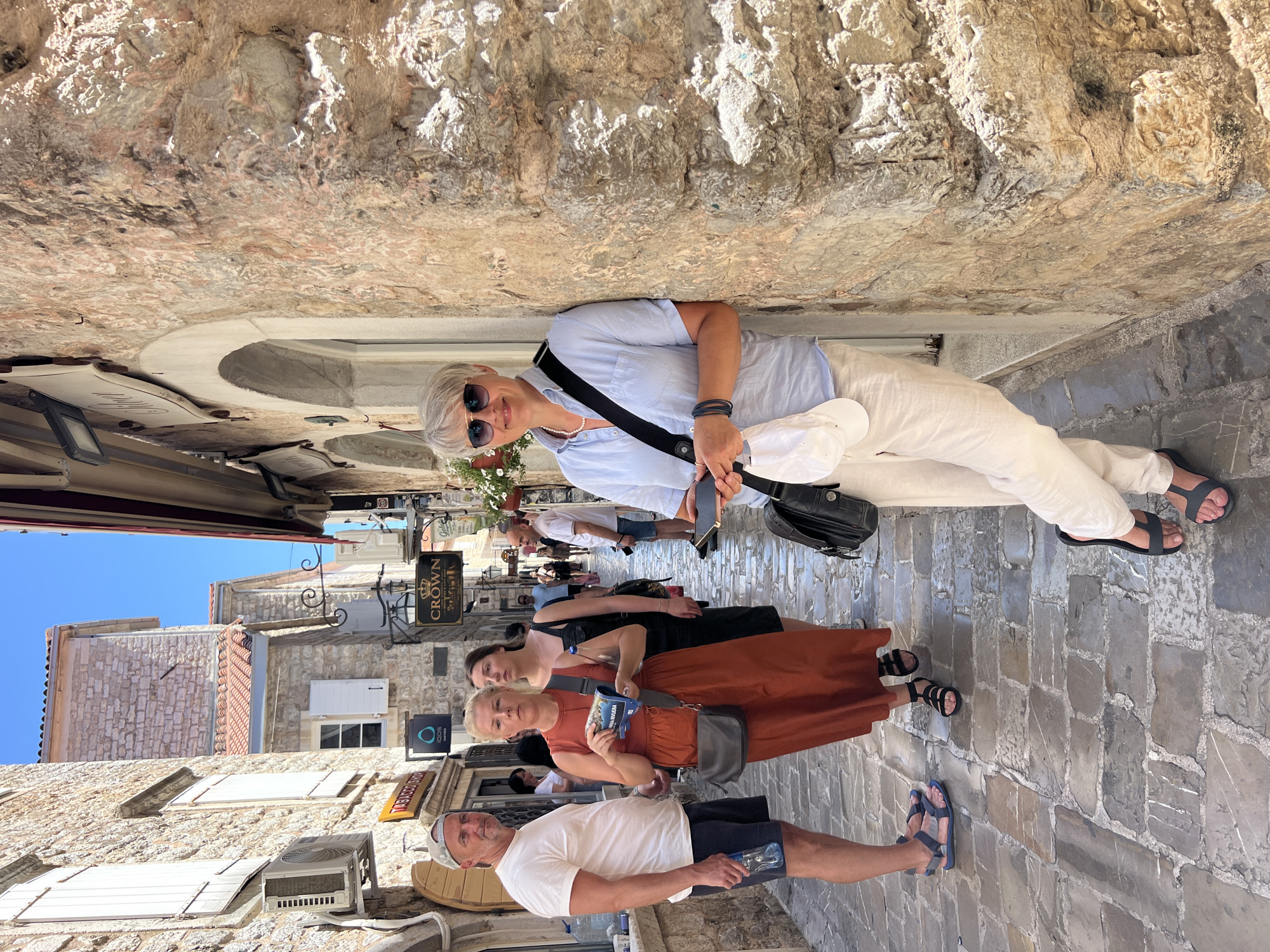 The image shows a group of people walking down a narrow, cobblestone street in Budva, Montenegro. The buildings on either side of the street are made of stone, and some have red tile roofs. The sky is blue and clear, suggesting it is daytime and the weather is sunny.
In the foreground, a woman with short gray hair and sunglasses is smiling at the camera. She is wearing a light blue short-sleeved shirt and white pants, with a black crossbody bag. Behind her, three other people are walking: a woman in a black dress and a rust-colored dress, holding a blue package, and a man in a white t-shirt and dark shorts. Further down the street, other people are visible, and shops with signs are lining the sides. One sign clearly reads "CROWN". The atmosphere appears casual and relaxed, suggesting a tourist or leisure activity.