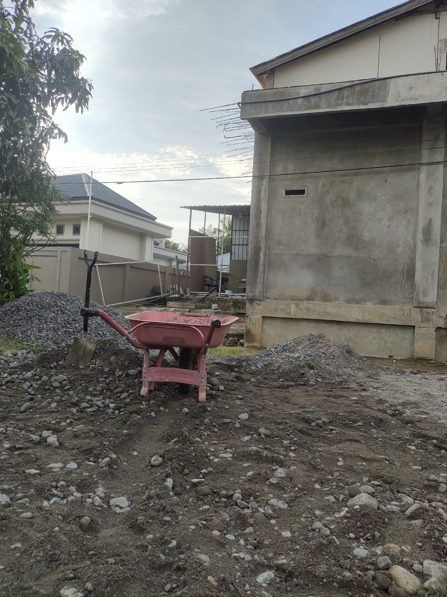 In an outdoor construction site in Meunasahaluedrienlhoksukon Barat, Indonesia, a red wheelbarrow with a shovel stuck in the dirt next to it sits in the foreground amidst piles of gravel and soil. A large, unfinished concrete building with exposed rebar dominates the right side of the image, while a smaller, modern house with a dark tiled roof is visible in the background to the left. Overhead, power lines crisscross the cloudy sky. The scene suggests ongoing construction work, with materials like gravel and soil readily available.