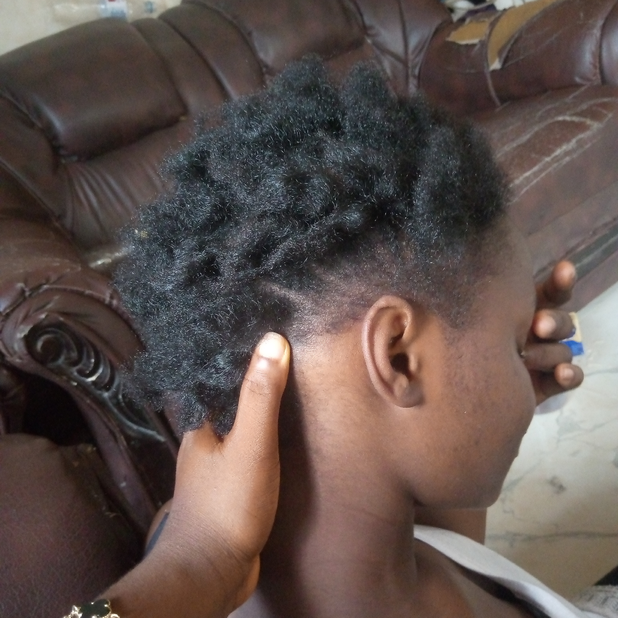 The image shows a person from their right side and back, focusing on their head and hairstyle. The person has short, black, coily or curly hair on the top of their head. The sides and back of the head are closely shaved or faded, with a distinct, curved line pattern shaved into the hair just above the ear. A hand with a dark skin tone is visible, gently touching the person's neck and the lower part of their head. This hand wears a gold-colored bracelet with black square elements on the wrist. The person is wearing a white garment, possibly a tank top or vest, visible around their shoulder and neck.
In the background, there is a large, brown upholstered sofa, which appears to be made of leather or faux-leather. The sofa shows visible signs of wear and tear, with some areas of the material appearing peeled or damaged, particularly towards the upper right side of the image. The setting appears to be indoors, likely a casual living space. The activity depicted suggests either hair styling, a head massage, or a comforting touch. There is no visible text in the image.
