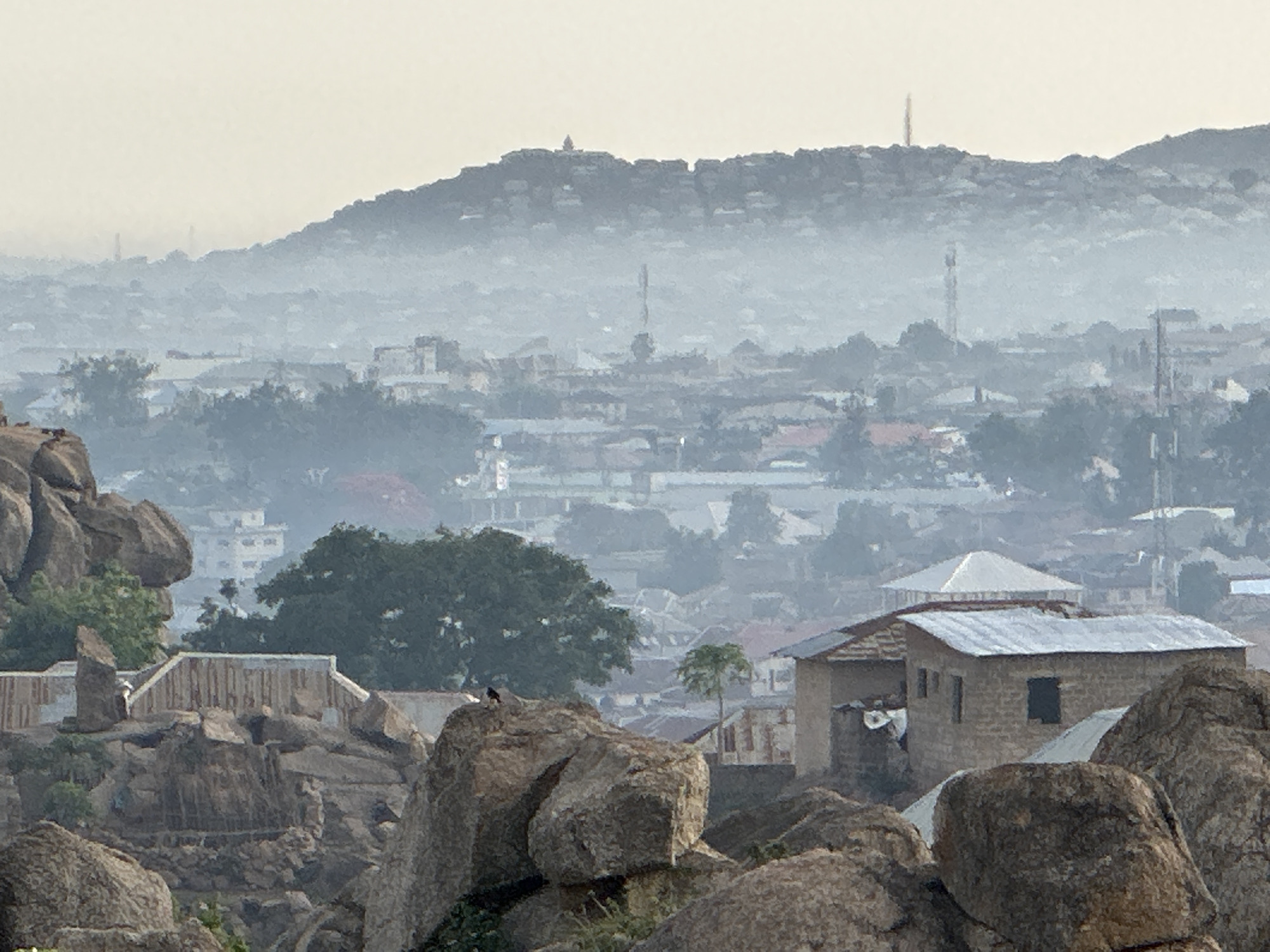 A hazy, panoramic view of Jos, Nigeria. In the foreground are large, rounded rocks. Several low-rise buildings with corrugated metal roofs are nestled amongst the rocks. In the middle ground, a cityscape unfolds, with more buildings, trees, and hints of infrastructure. The background consists of a hazy mountain range, with radio and cellphone towers dotting the landscape.