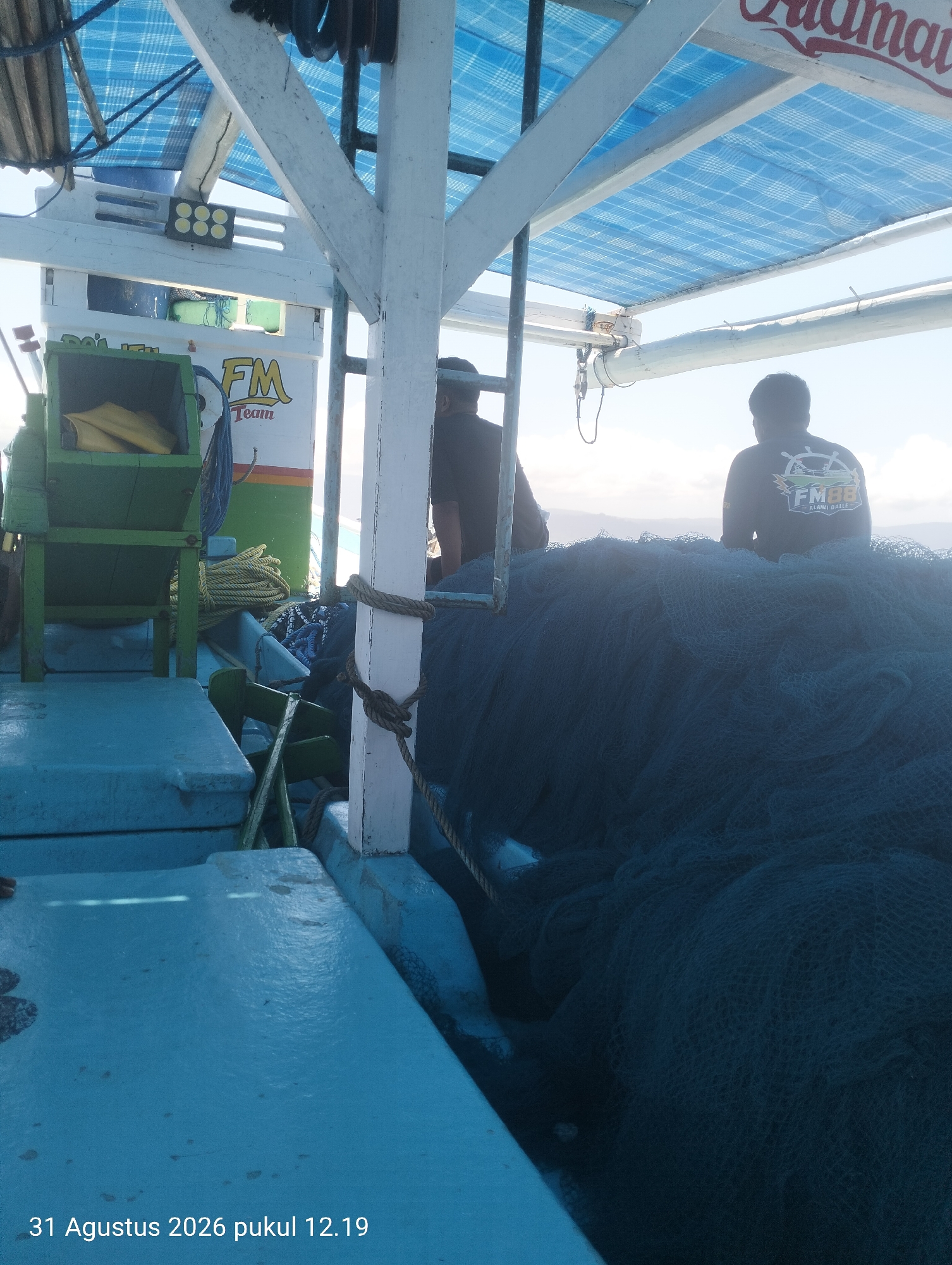 On August 31, 2026, at 12:19 PM in Pihingtado, Indonesia, two individuals are on a boat. The person on the left, facing away from the camera, is partially obscured by fishing nets. The person on the right, also facing away, wears a dark shirt with a logo that includes the text "FM88 ALAMAI DALLE". A large pile of dark blue fishing nets occupies the right side of the boat. In the background, a blue tarp covers the upper portion of the boat, supported by white beams. Green fishing equipment, including a net winder and coiled yellow rope, is visible on the left side of the boat. The boat appears to be at sea, with a hazy horizon and sky in the background.