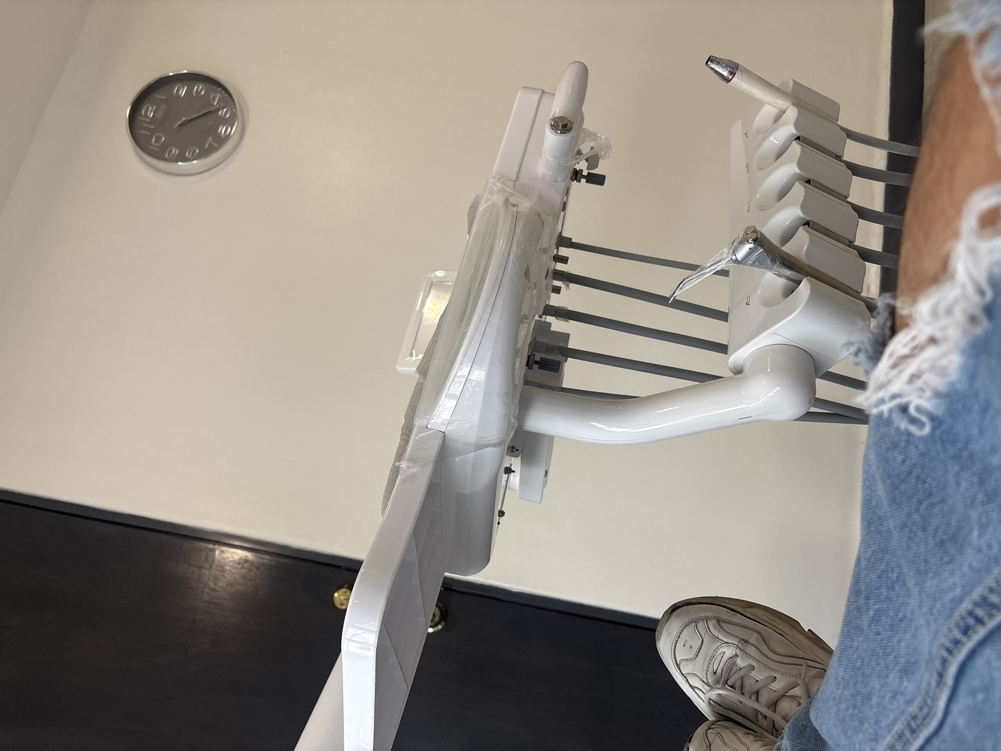 The image depicts a dental operating room environment. Dominating the foreground is a dental chair and equipment arm, both in white and partially wrapped in plastic, suggesting a new or recently cleaned setup. The chair and arm are positioned against a cream-colored wall, upon which hangs a round clock indicating the time is approximately 11:11. Below, a dark-colored wainscoting or cabinet runs along the lower portion of the wall. The lower part of a person is visible on the right side of the image, wearing jeans and a white sneaker. The scene suggests a dental clinic or office, with the focus on the equipment prepared for a patient. This particular location is not confirmed as Puebla, Mexico, based solely on the visual content of the image.