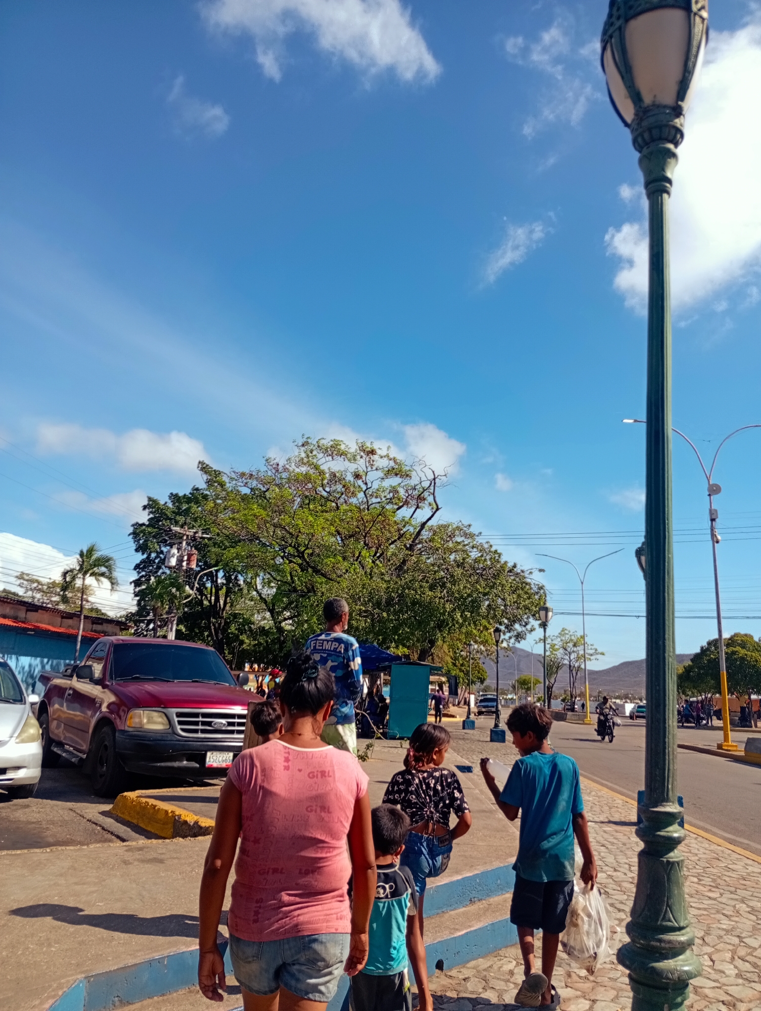 The image depicts a street scene in Juan Griego, Venezuela, on a sunny day with a bright blue sky and scattered white clouds. In the foreground, a woman wearing a pink t-shirt and denim shorts walks away from the camera, her back to the viewer. Two children are walking with her, a boy in a blue t-shirt and shorts carrying a plastic bag, and another child in a blue and black shirt. Behind them, a red pickup truck is parked on the side of the road. To the right of the scene, a decorative lamppost with a green base and ornate lighting fixture stands prominently. A paved road runs parallel to the sidewalk, with vehicles and people visible in the distance. Trees with green foliage line the street, and mountains can be seen in the background. The overall impression is a snapshot of everyday life in a Venezuelan town.