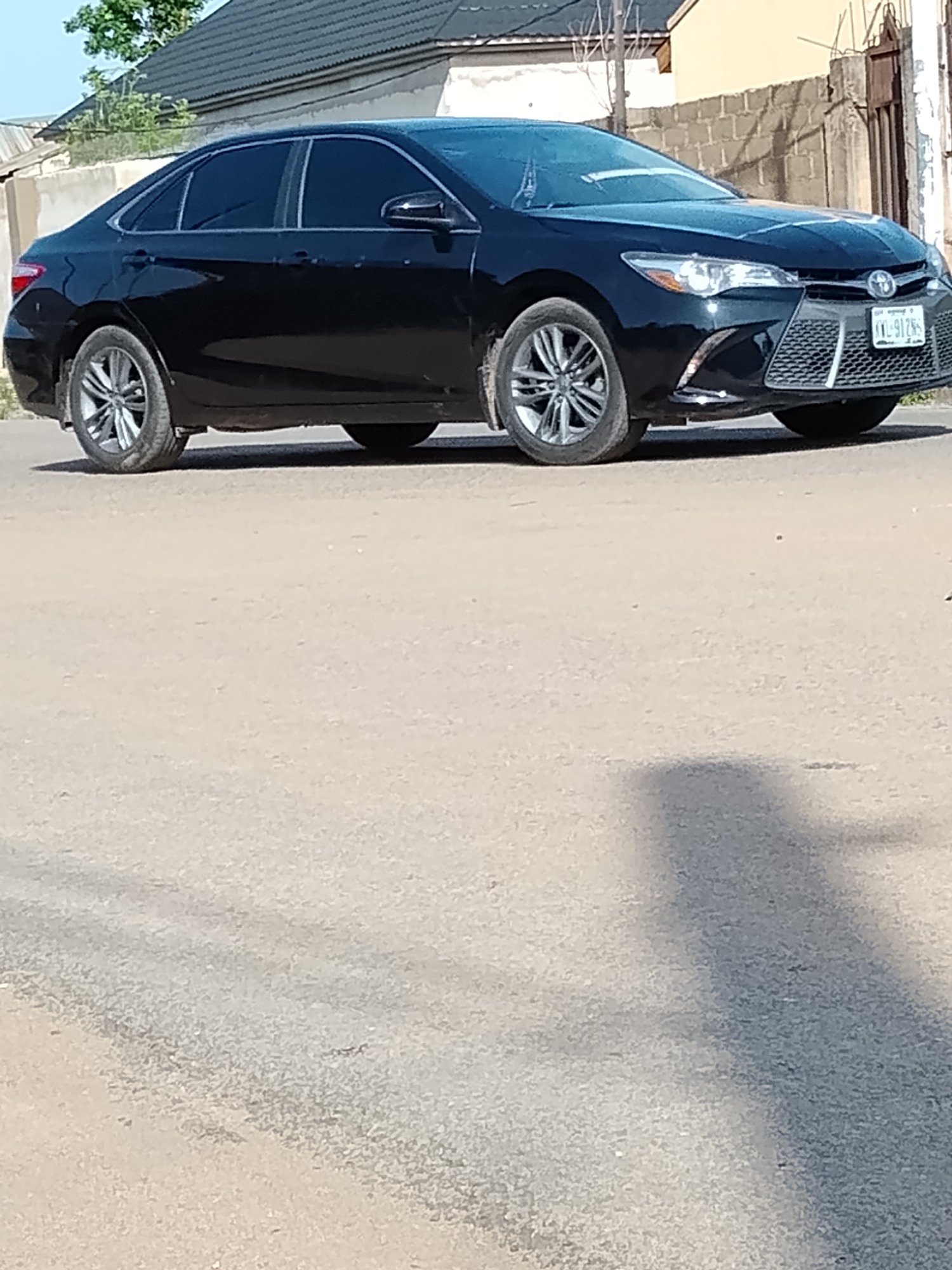 A black Toyota Camry sedan is parked on a paved road in Wauro Leggal, Nigeria. The car is positioned at an angle, facing towards the right of the frame. It has tinted windows and a license plate that reads "XXL-9125S". The vehicle is in the foreground, with a blurry background that includes buildings and some vegetation. The lighting suggests it is daytime, and a shadow is cast on the road to the right of the car, indicating the presence of an unseen object or person.