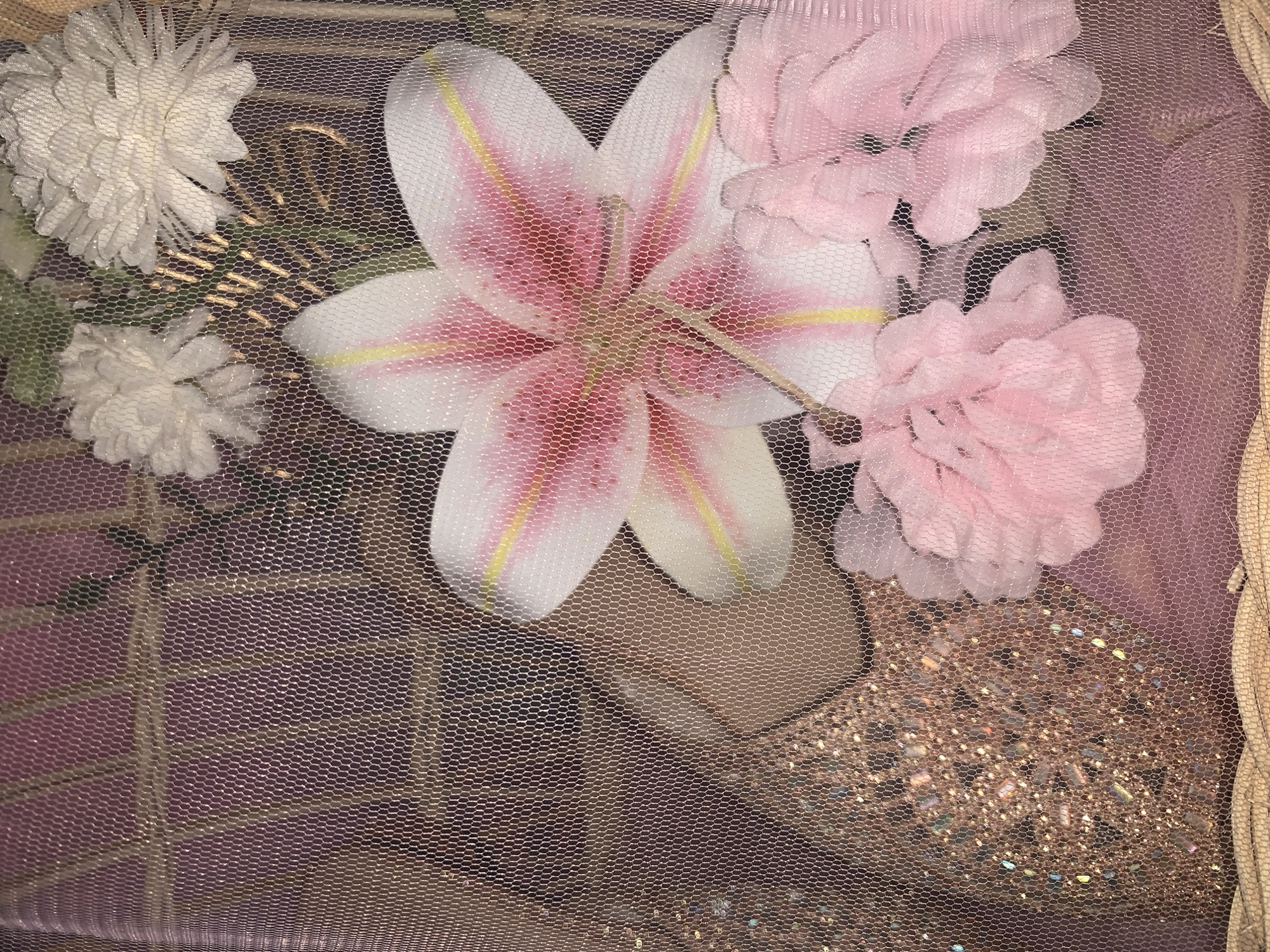 The image depicts a close-up view of artificial floral arrangements and a decorative, sparkly object, possibly a bauble, all contained within a netted or screened enclosure. The main subjects are a large pink and white lily, several smaller pink flowers, and two white, ruffled flowers at the top. A rose gold, faceted, spherical object with glitter is visible on the left. The background appears to be a textured, possibly fabric, surface in shades of purple and pink. The netting has a hexagonal pattern and casts a faint shadow. There are no people or animals present. The lighting is soft, creating a gentle glow and highlighting the textures and sparkle of the objects. The scene does not contain any identifiable elements that would place it specifically in Kuala Pembuang, Indonesia.