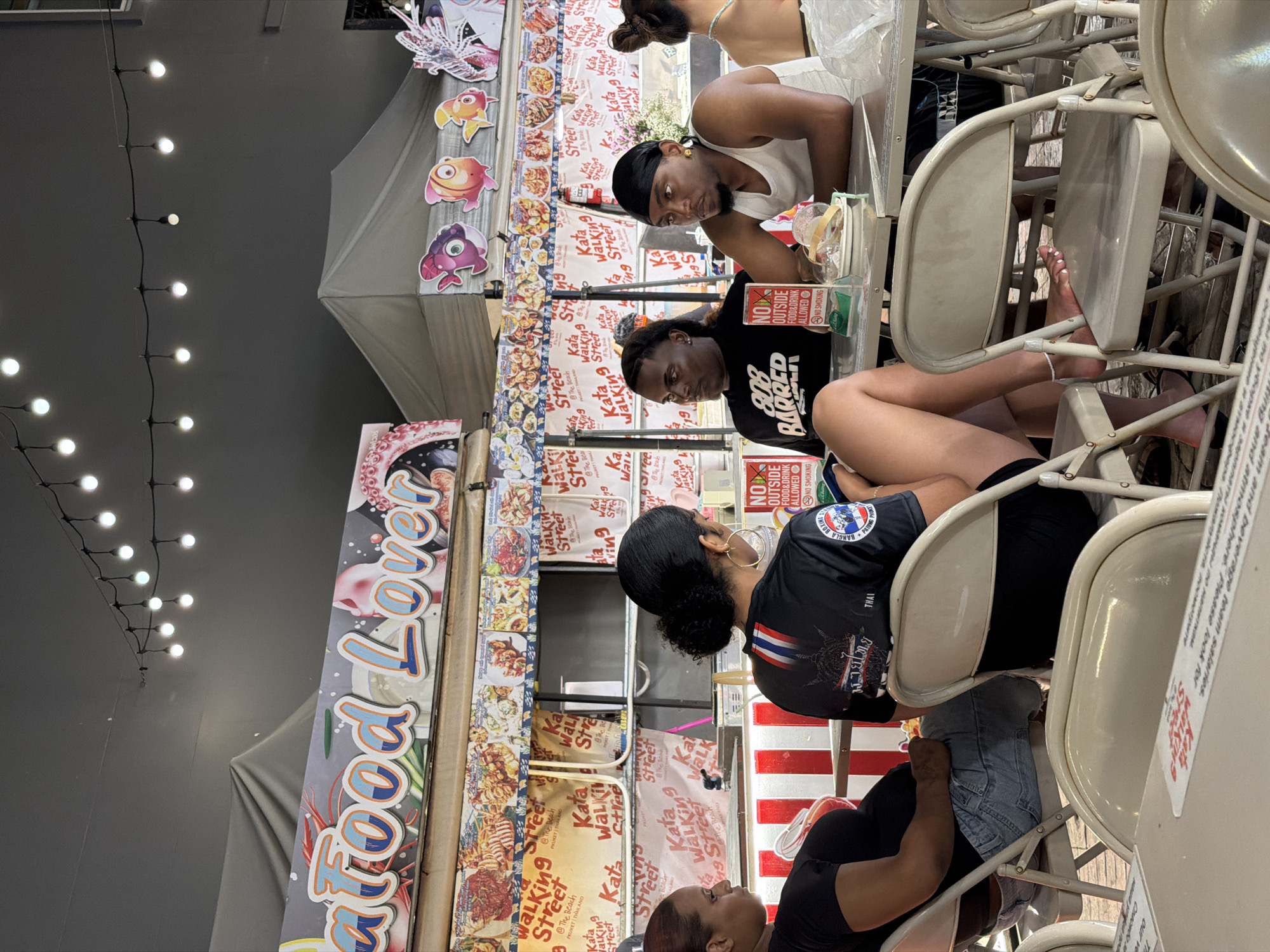 The image captures a scene at a street food stall in Karon, Thailand, with a sign prominently displaying "Seafood Lover." The stall is adorned with decorative fish cutouts and strings of lights. In the foreground, several people are seated at tables, engaged in conversation and dining.
A woman with dark curly hair, wearing a black t-shirt with a Thai flag design and large hoop earrings, is seated at a table. Next to her, a man wearing a black t-shirt with "808 BARRED" printed on it, and another man in a white tank top and black durag are also seated. The man in the white tank top has a small plate with food in front of him. In the background, another woman with dark hair is visible, seated at a table.
The atmosphere appears casual and lively, characteristic of an outdoor dining establishment in a tourist area. The presence of multiple food menus and the general layout suggest a bustling food court or market. The lighting indicates it might be daytime or early evening.