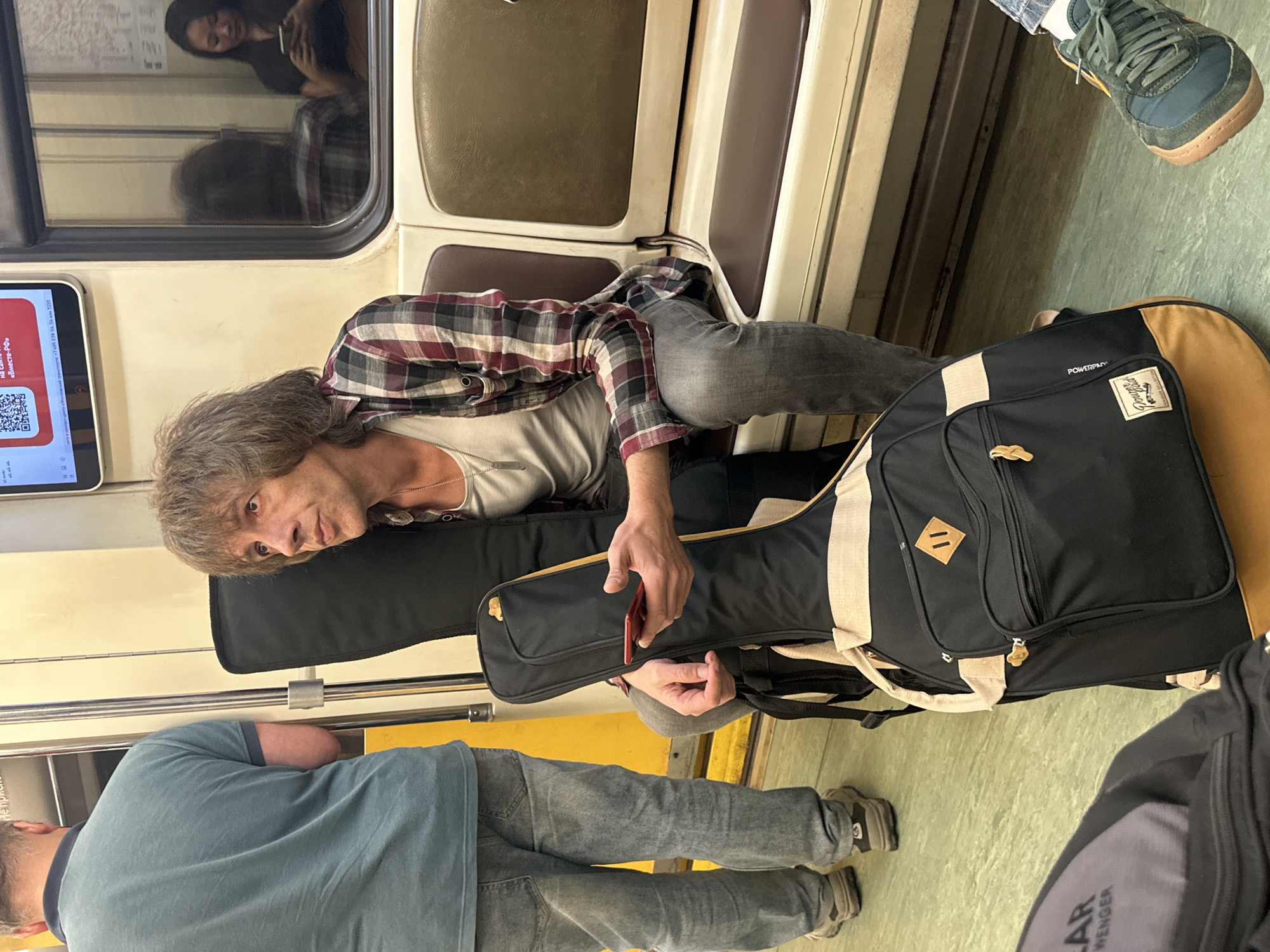 A man with shaggy brown hair and a plaid shirt sits on a subway seat in Moscow, Russia. He holds a red smartphone in his right hand, which rests on a black and tan guitar case. He is looking to his left, out of frame. Behind him, another passenger, a woman, is visible in the reflection of the window, looking at her phone. The subway car is illuminated by artificial light. The floor is a light green linoleum, and the seats are upholstered in a brown material. A man in a blue t-shirt and jeans stands to the left of the frame, facing away from the camera.