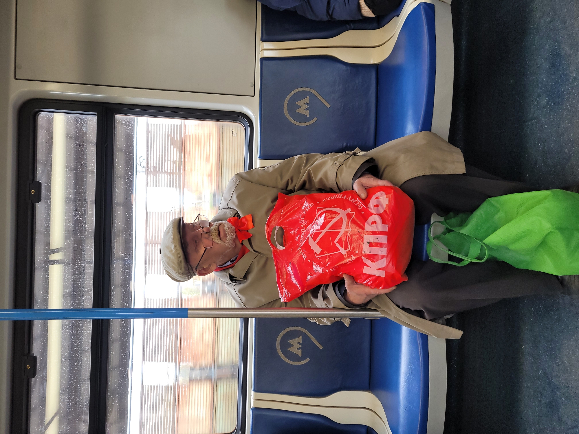 An elderly man wearing glasses, a hat, a tan jacket, and an orange scarf sits on a blue subway seat in what appears to be a train car, holding a red plastic bag with the image of a hammer and sickle, and Cyrillic text that appears to be "KPRF." A green reusable shopping bag is also on the seat next to him. The subway train is identified by the “M” symbol on the seat as being located in Moscow, Russia.