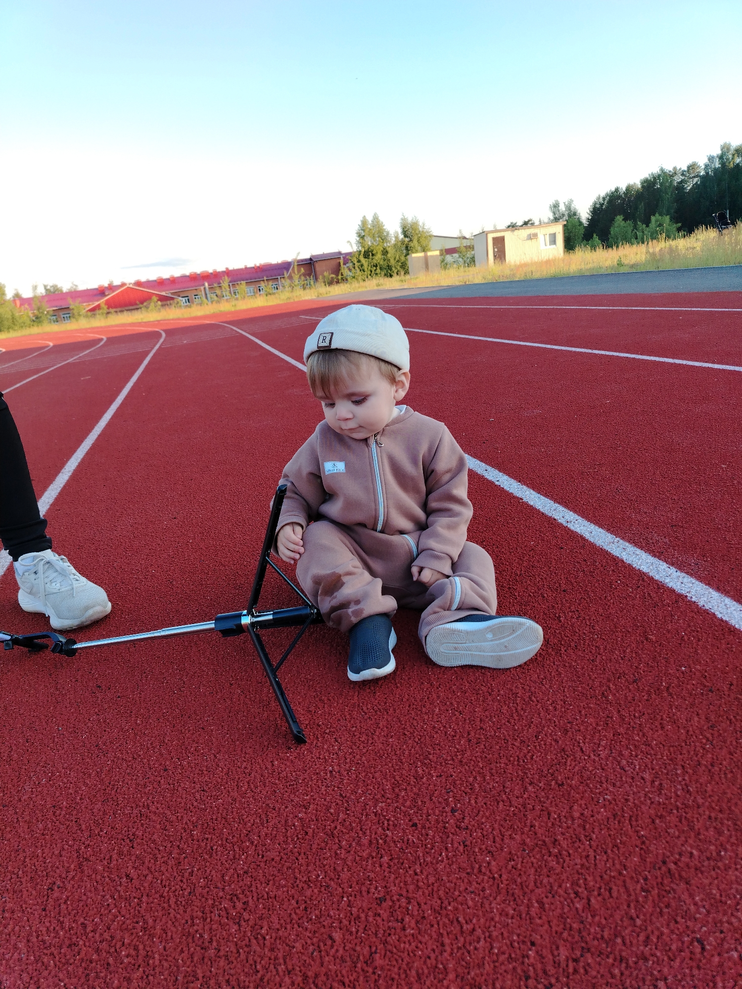 A young child sits on a red athletic track with white lane markings, looking down at a small black and silver tripod-like device (possibly a selfie stick). The child wears a beige/brown full-body tracksuit and a light-colored beanie with a black patch featuring a white "R". On the tracksuit, a small rectangular patch bears the letters "RK" and some indistinct text below it. Part of an adult's leg and foot, clad in black pants and white sneakers, is visible to the left of the child and the device.
The scene is outdoors, under a clear, bright sky, suggesting late afternoon or early evening. In the background, buildings with red roofs are visible, along with green trees and what appears to be a smaller, light-colored shed-like structure. A strip of gray asphalt or gravel separates the track from the grassy/wooded area. The setting is a sports ground or stadium, located in Noviy, Russia. The child appears calm and focused on the object in front of them.