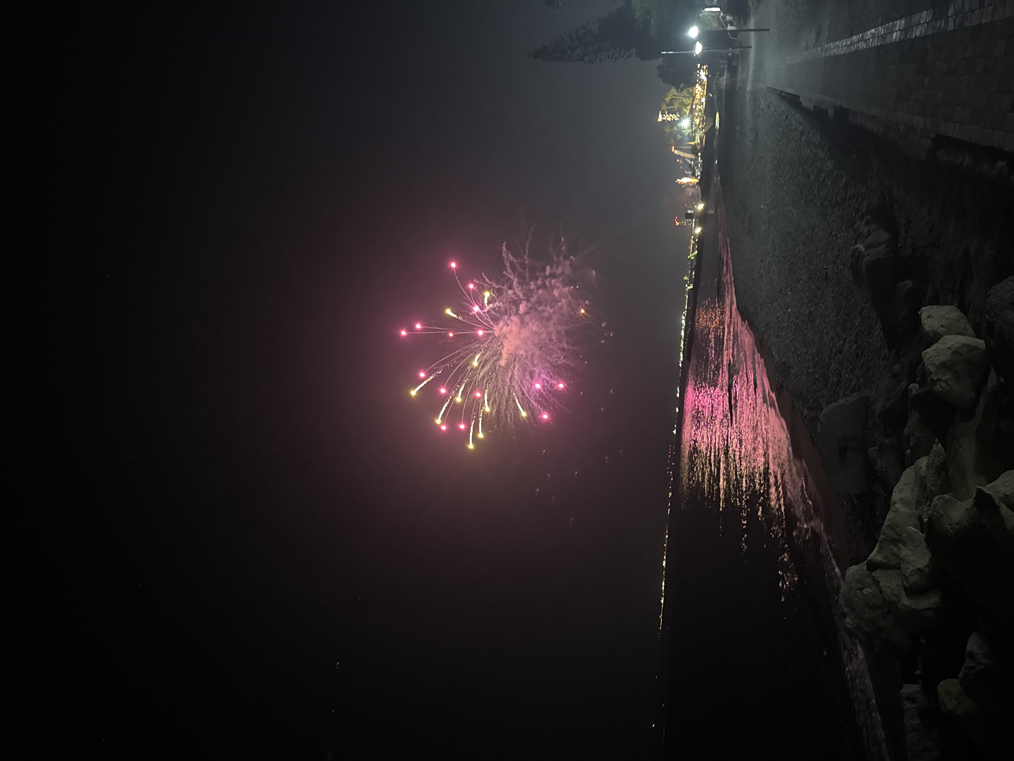 The image captures a nighttime scene on the coast of Agios Tychona Community, Cyprus, featuring a vibrant fireworks display over the sea. The fireworks, predominantly pink and yellow, illuminate the dark sky, with some reflecting on the water's surface. The sea gently laps at a sandy and pebbly beach, with large rocks visible in the foreground. Along the coastline, a promenade is visible, lined with trees and lampposts, indicating an urban or resort area. Distant lights from buildings and structures suggest a populated coastline. The overall atmosphere is festive and celebratory, likely during an event or holiday.