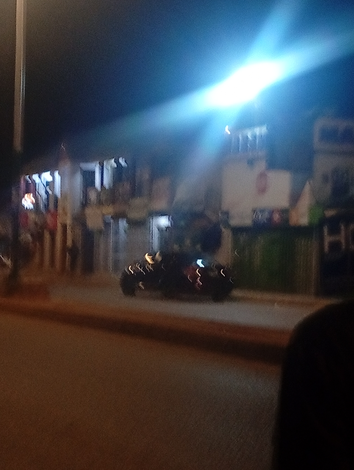 The image depicts a blurry nighttime street scene in Bukuru, Nigeria. In the foreground, a dark shape, possibly a vehicle, is visible with illuminated streaks suggesting motion. The background features buildings with some illuminated windows and what appears to be a bright light source dominating the upper portion of the frame. The overall impression is one of a busy, albeit dimly lit, urban environment at night, with a focus on the movement and artificial lighting that characterizes such settings.