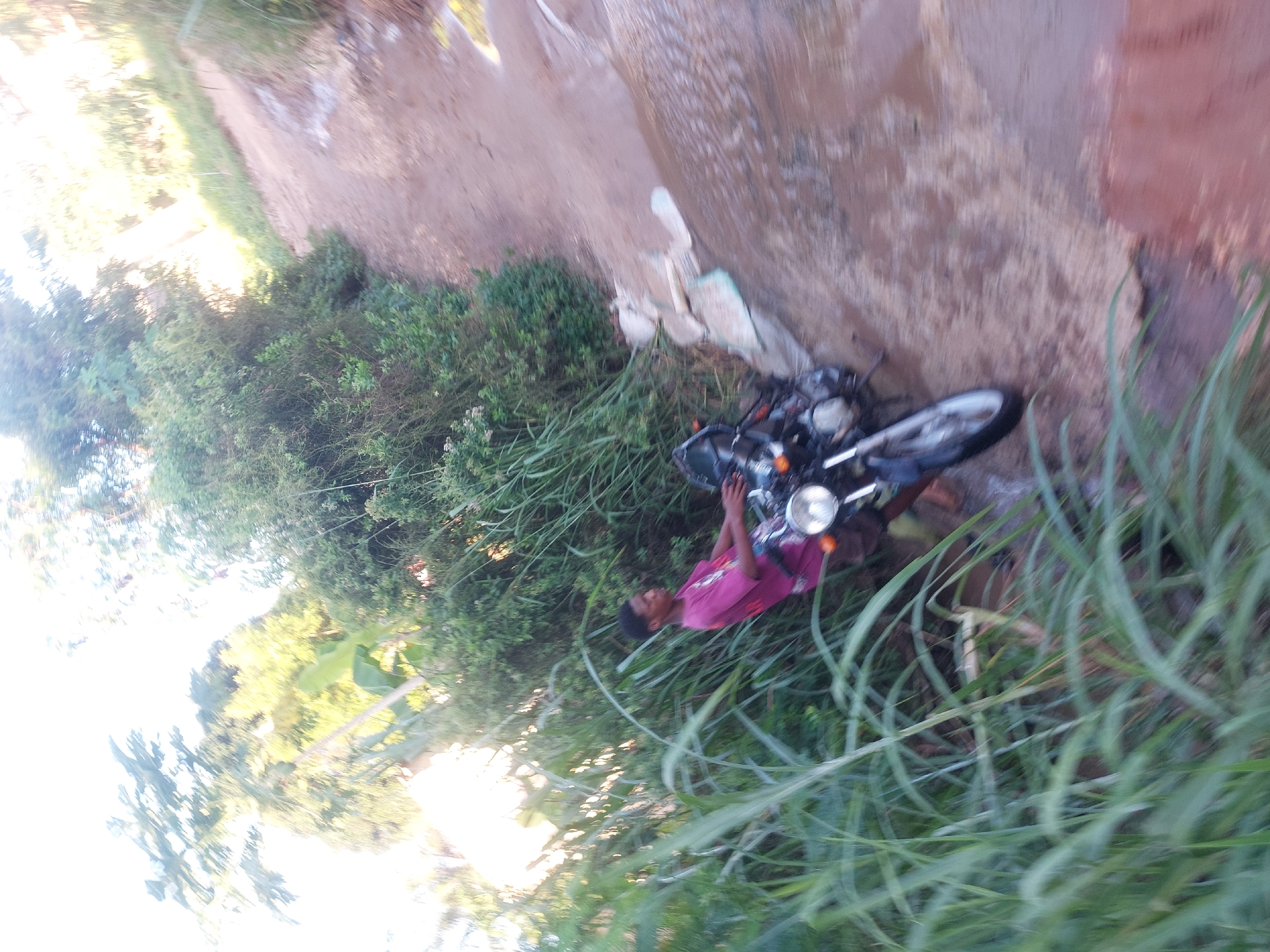 The image depicts a man wearing a pink shirt standing next to a black motorcycle on the side of a dirt road. The motorcycle is partially obscured by tall green grass and vegetation. The road runs along a raised bank, and there is additional green vegetation, including trees and shrubs, visible in the background. The location is likely in a more rural or undeveloped area, given the dirt road and surrounding vegetation. Without additional location information, it is impossible to determine the specific area within Dar es Salaam.