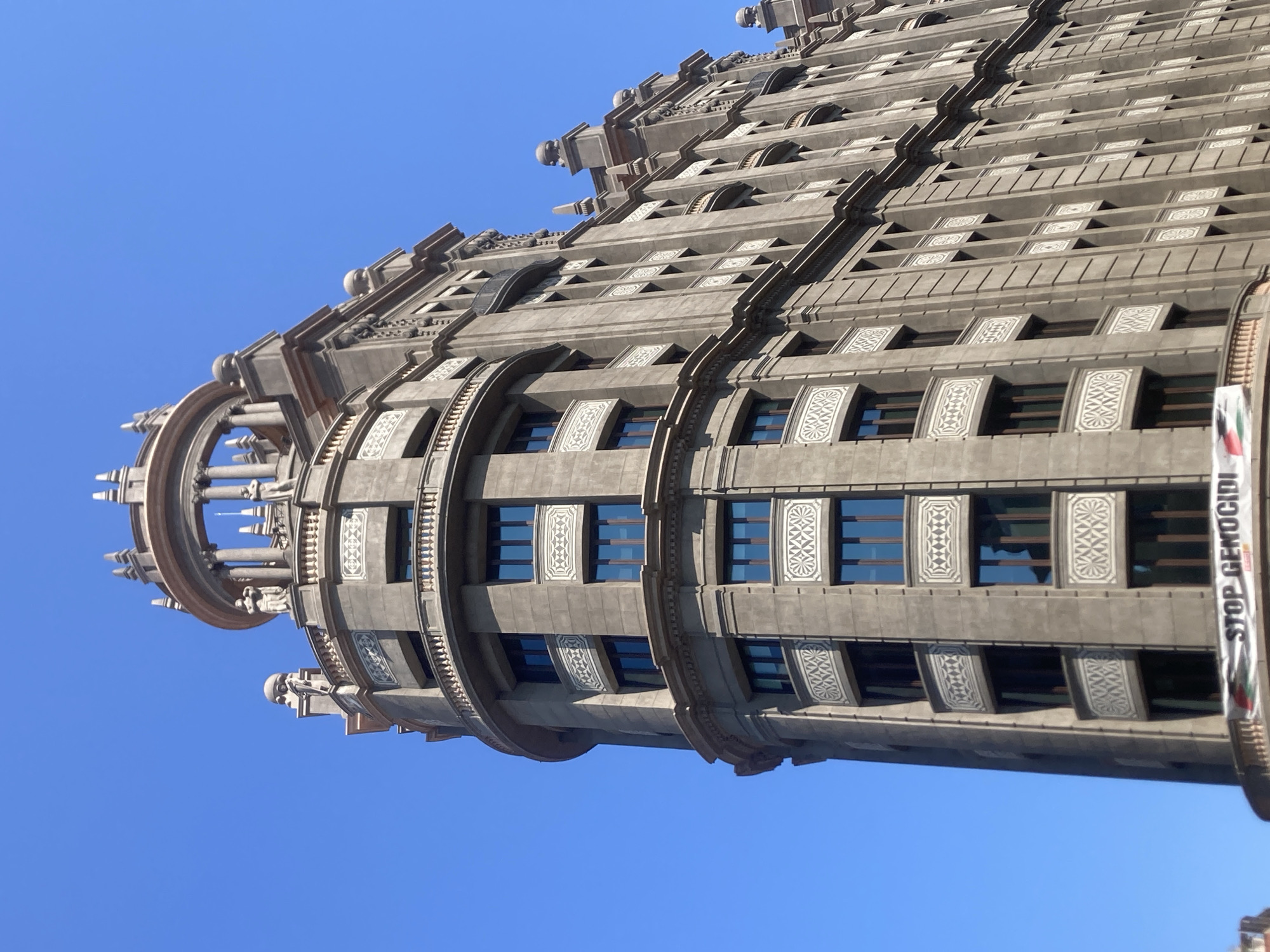 The image shows a close-up, low-angle shot of a tall, ornate building against a clear blue sky. The building is constructed of a light-colored stone and features a curved facade on the left, with multiple stories of windows and decorative architectural elements. The upper portion of the building culminates in a domed structure with columns and small statues. On the lower part of the building, a banner is visible with the text "STOP GENOCIDI" written in white letters on a dark background, with some colorful bands to the left of the text. The building appears to be located in Barcelona, Spain, based on the distinctive architectural style which is characteristic of the city. The scene is a street-level view, with no other buildings clearly visible in the immediate vicinity, suggesting an urban setting. The lighting indicates it is daytime, with the sun casting shadows on the building's details.