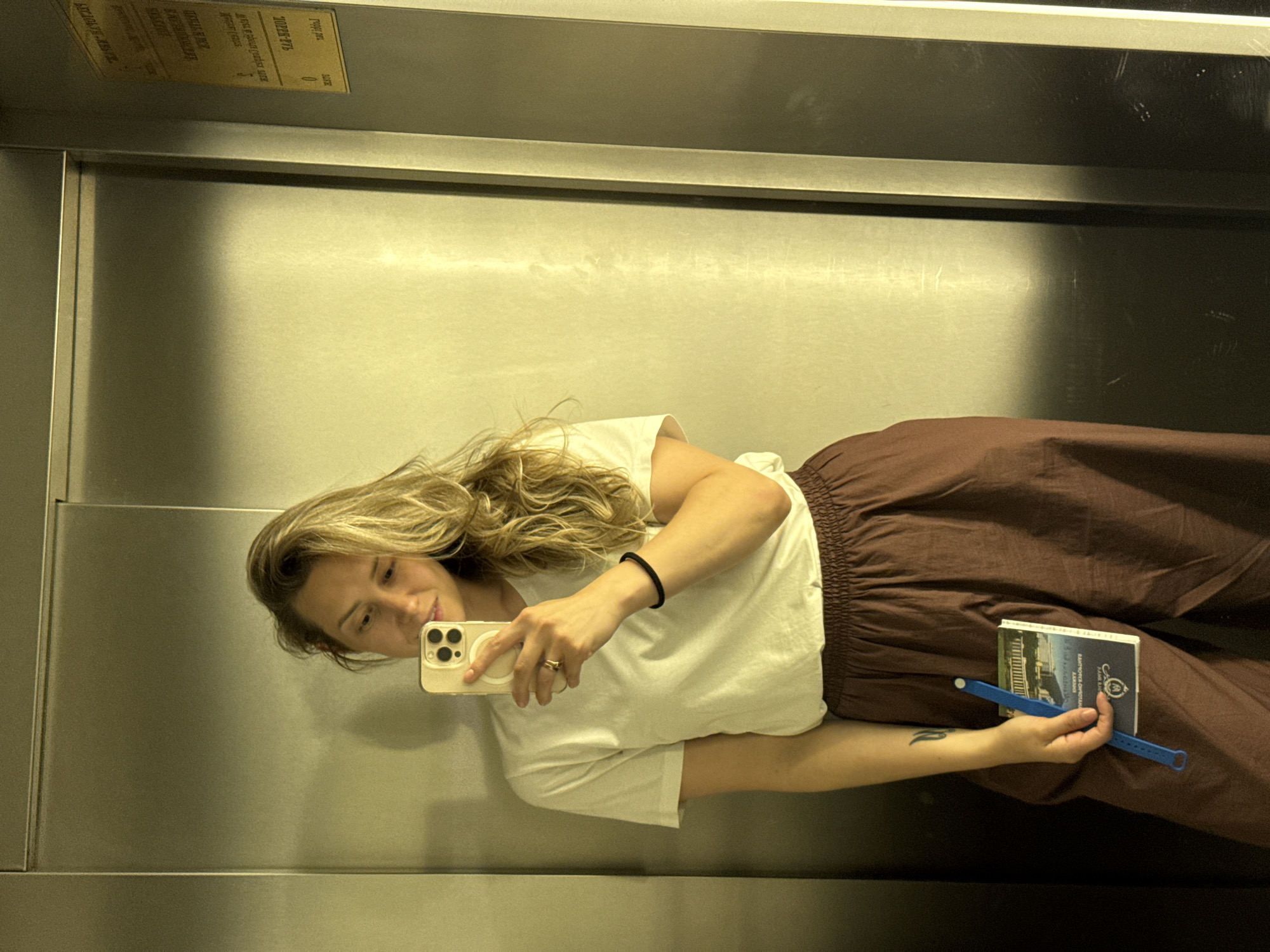 A woman with blonde, wavy hair is taking a selfie in an elevator. She is wearing a white t-shirt, maroon pants, and a black bracelet on her wrist. She is holding a phone in her right hand and a small booklet and a blue pen in her left hand. A tattoo is visible on her left arm. The elevator walls are made of brushed metal, and there is a yellow sign with text on the right side of the elevator. The scene appears to be set in the city of Nebug, Russia.