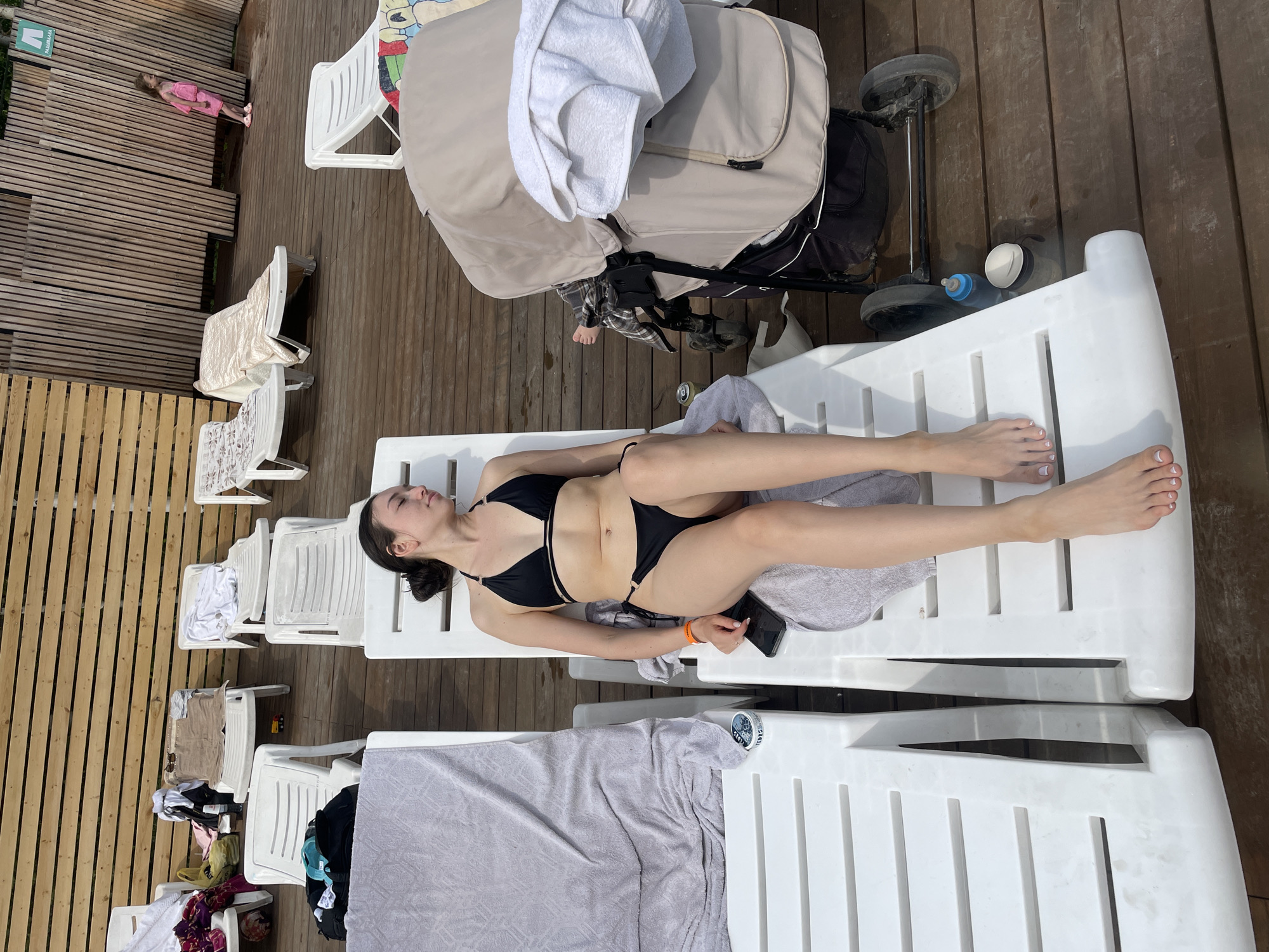 A woman in a black bikini is sunbathing on a white lounge chair. She is lying on her back with her eyes closed, her face tilted upwards towards the sun. Her legs are extended and she is holding a smartphone in her right hand. Another lounge chair is visible in front of her, and beyond that, a baby stroller. In the background, a young girl in a pink outfit stands on a wooden deck. The scene appears to be outdoors, possibly at a resort or poolside. The time of day seems to be daytime, with bright sunlight casting shadows. The location is identified as Kazan, Russia.