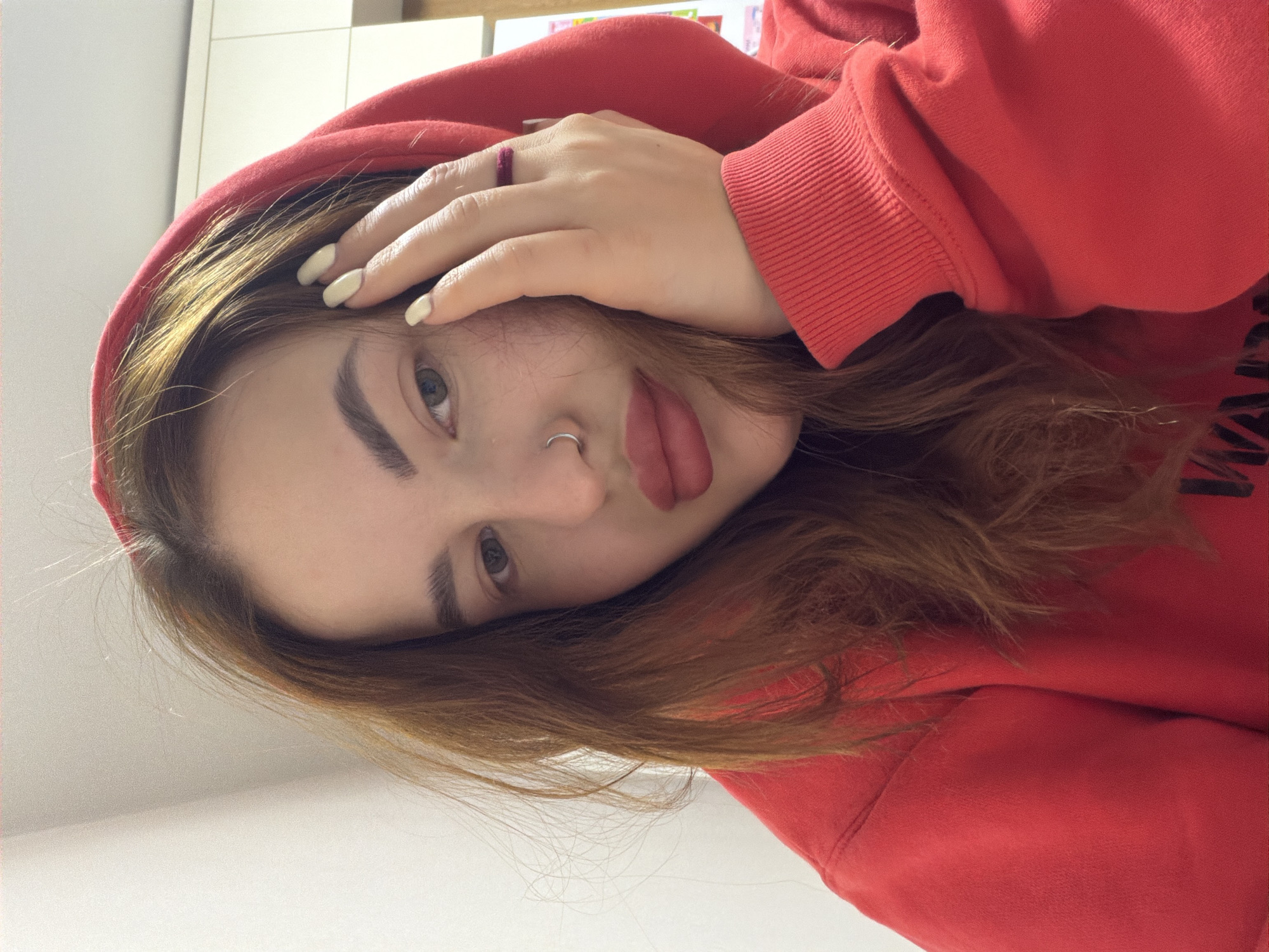 A young woman is depicted in a close-up, portrait-style photograph, likely a selfie, taken indoors in Krasnodar, Russia. She has long, reddish-brown hair, prominent dark eyebrows, and light-colored eyes that gaze directly at the camera. She is wearing a vibrant red hooded sweatshirt, with the hood up, framing her face. A small, silver hoop piercing is visible in her septum, and her lips are painted a reddish-pink color.
Her left hand is positioned across her forehead, partially obscuring her right eye. Her fingernails are manicured and painted a creamy off-white shade, and a dark red ring is worn on one of her fingers. The background is plain and light-colored, suggesting a simple indoor environment like a wall or interior surface. The lighting appears soft and natural, illuminating her features. Her expression is calm and direct, conveying a casual and perhaps slightly contemplative mood. No discernible text or specific location markers are visible within the image itself.