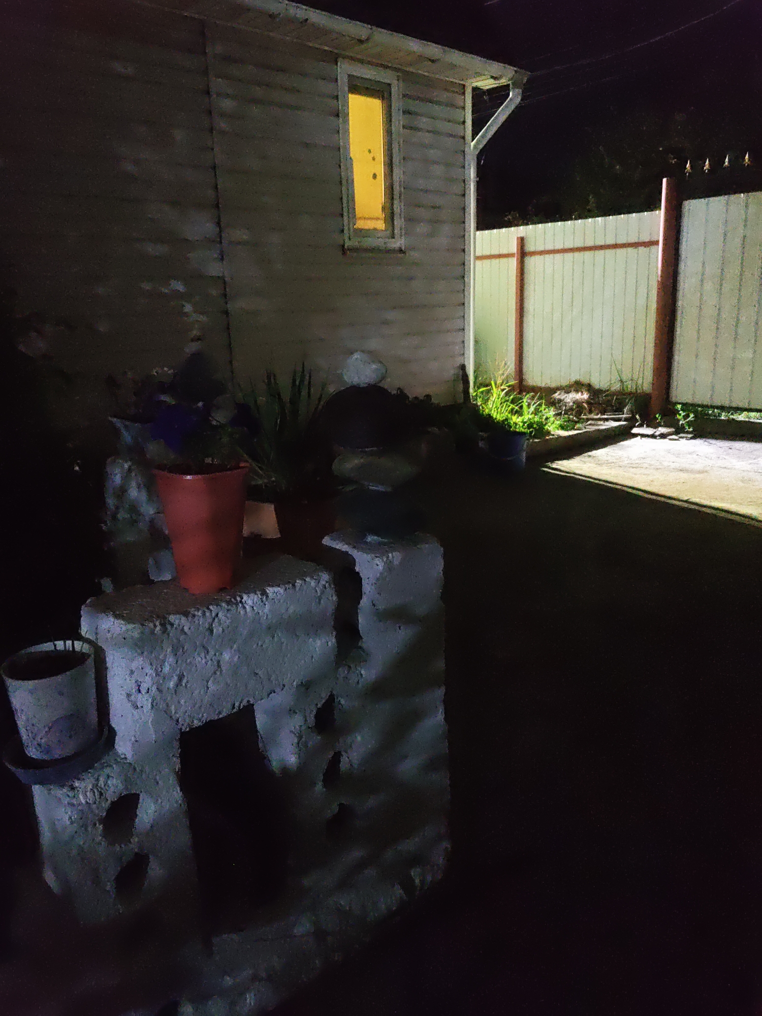 The image depicts a dark backyard scene at night. On the left, a dilapidated wooden building with a small, illuminated window is visible. In the foreground, there are several concrete blocks arranged to form a makeshift structure, with a red flower pot and another grey pot on top. Some greenery and other objects are scattered around the area. To the right, a light-colored fence with wooden posts separates the yard from an unseen area. The ground appears to be asphalt or concrete. The overall impression is one of a neglected outdoor space, possibly in a residential area. There are no people or animals visible in the frame. The lighting suggests artificial light sources, possibly from inside the building or from external illumination. The specific location in Rostov-on-Don, Russia, cannot be determined from the visual information provided.