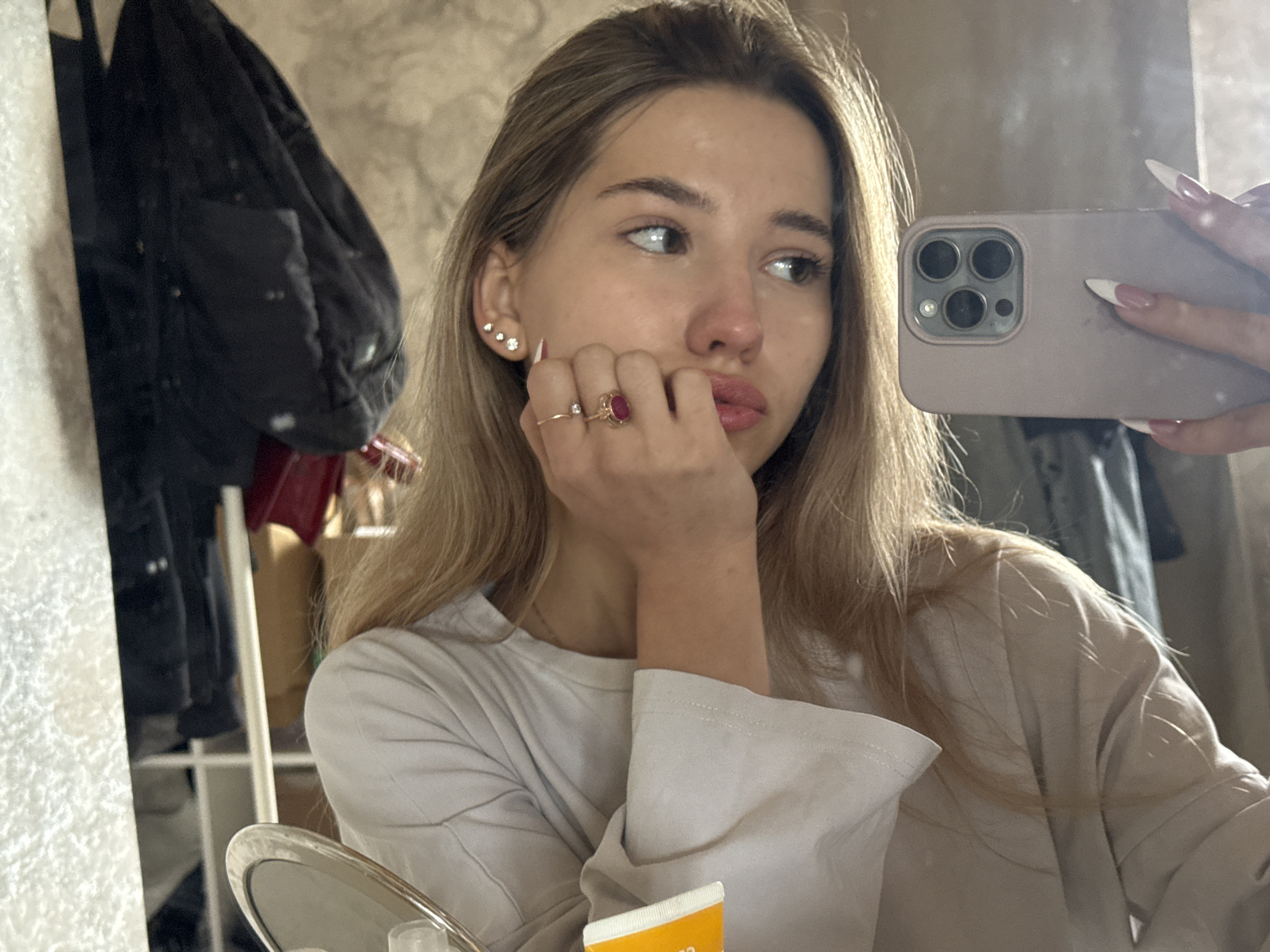 A young woman with blonde hair is taking a selfie in a room. She is wearing a light-colored, long-sleeved shirt and has rings on her fingers. Her nails are long and manicured. She is holding a phone up to her face, and the camera is visible. In the background, there's a rack with dark clothing, a mirror, and other household items. The location is not discernible as Noviy, Russia.
