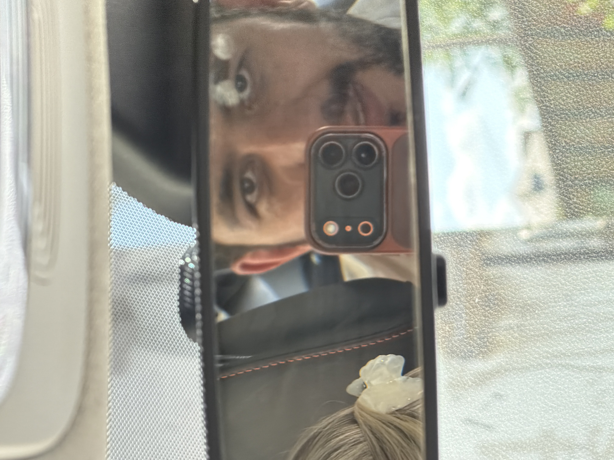 The image shows a reflection of a person and their phone in a car's rearview mirror. The person's face is visible, and they are holding up the phone with the camera facing the mirror. The reflection also shows the inside of the car and a glimpse of the outside environment, possibly a street or parking lot with trees or buildings. The background is blurry, suggesting the reflection is the main focus. The location cannot be identified as Krasnoyarsk, Russia.