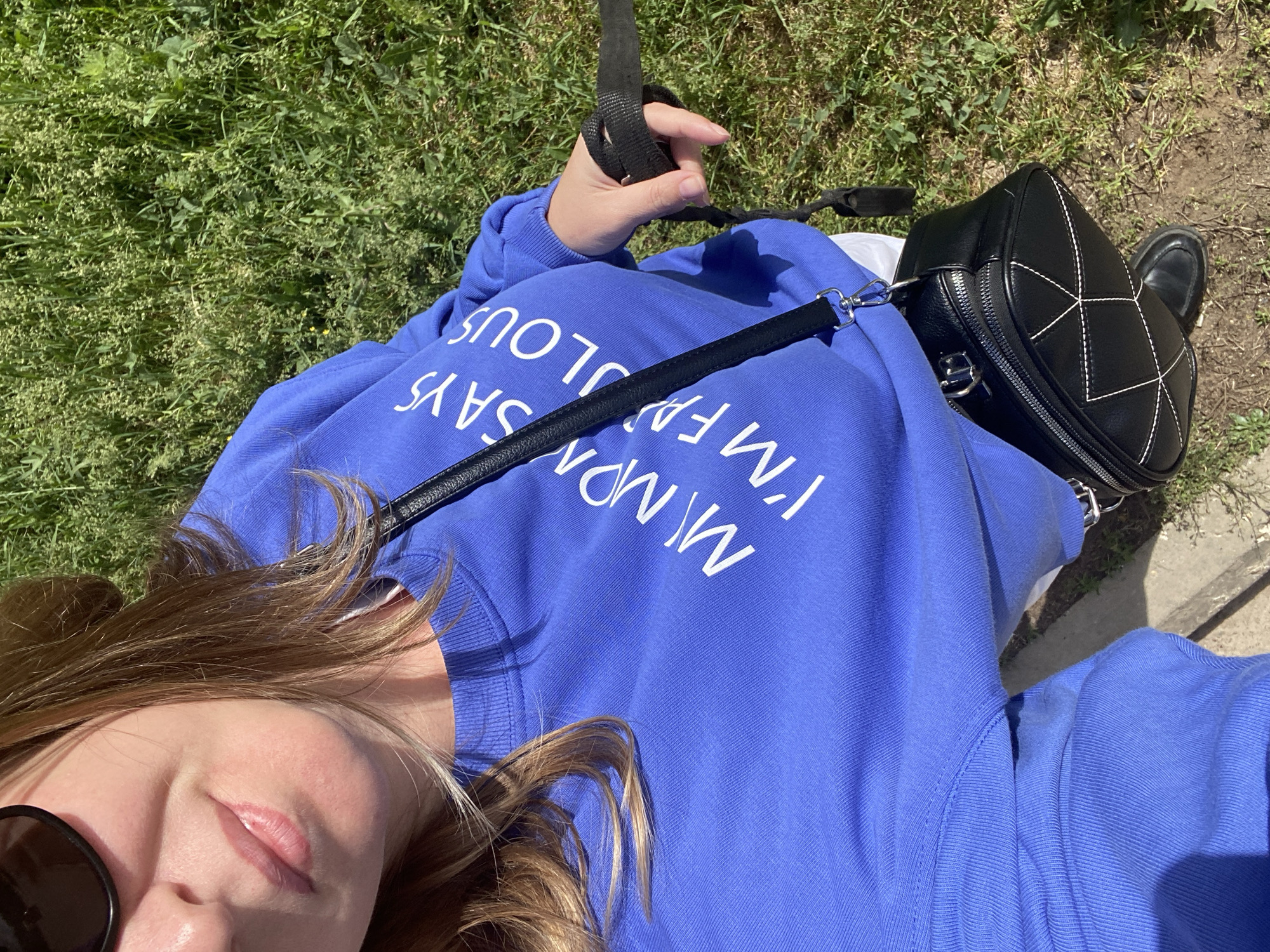 A woman lies on grass next to a paved surface. She wears a blue sweatshirt with white lettering, sunglasses, and carries a black purse. One hand holds a black strap.