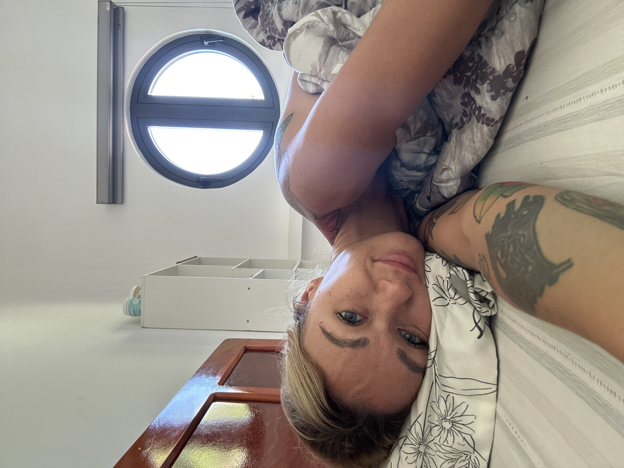 The image shows a woman lying down, seemingly in a bedroom. She has blonde hair and visible tattoos on her arms. She appears to be under a blanket or comforter, with a patterned pillow near her head. In the background, there is a round window and a white shelf. The room has a door with a dark wood frame and a shiny, reflective surface. It is not possible to pinpoint the exact location in Mũi Né, Vietnam without additional information.
