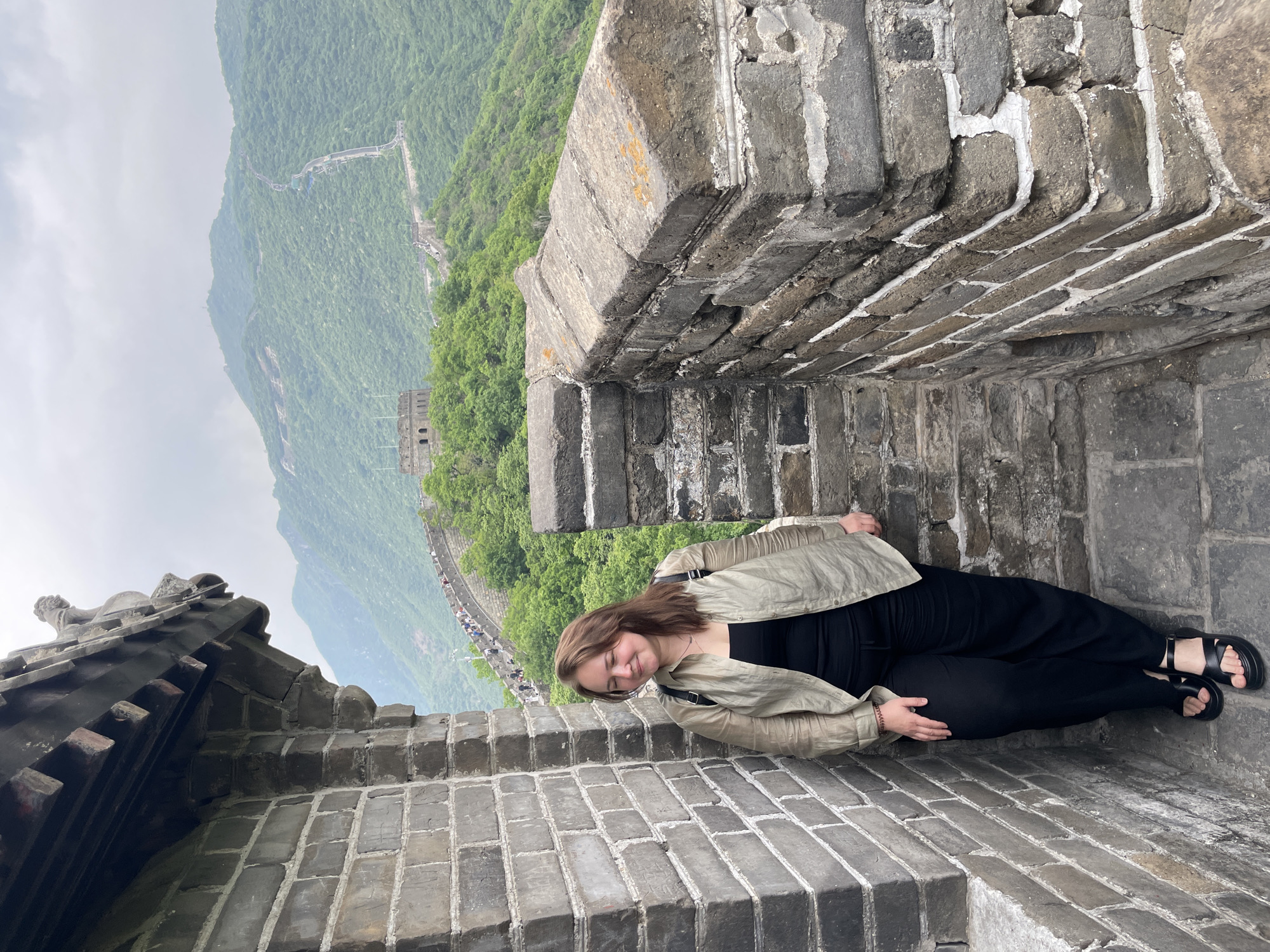A sideways, eye-level, medium shot depicts a woman lying on the Great Wall of China, in Huairou District, China. She faces the viewer and is lying on her right side, with her head near the bottom of the image and her feet near the top. She wears a light brown jacket over a black top and black pants, and black sandals. The woman's eyes are closed and she seems to be resting. She is framed by the stone wall of the Great Wall. In the background, a green mountain range stretches into the distance. A section of the Great Wall can be seen winding along the crest of the mountains. The sky is overcast.