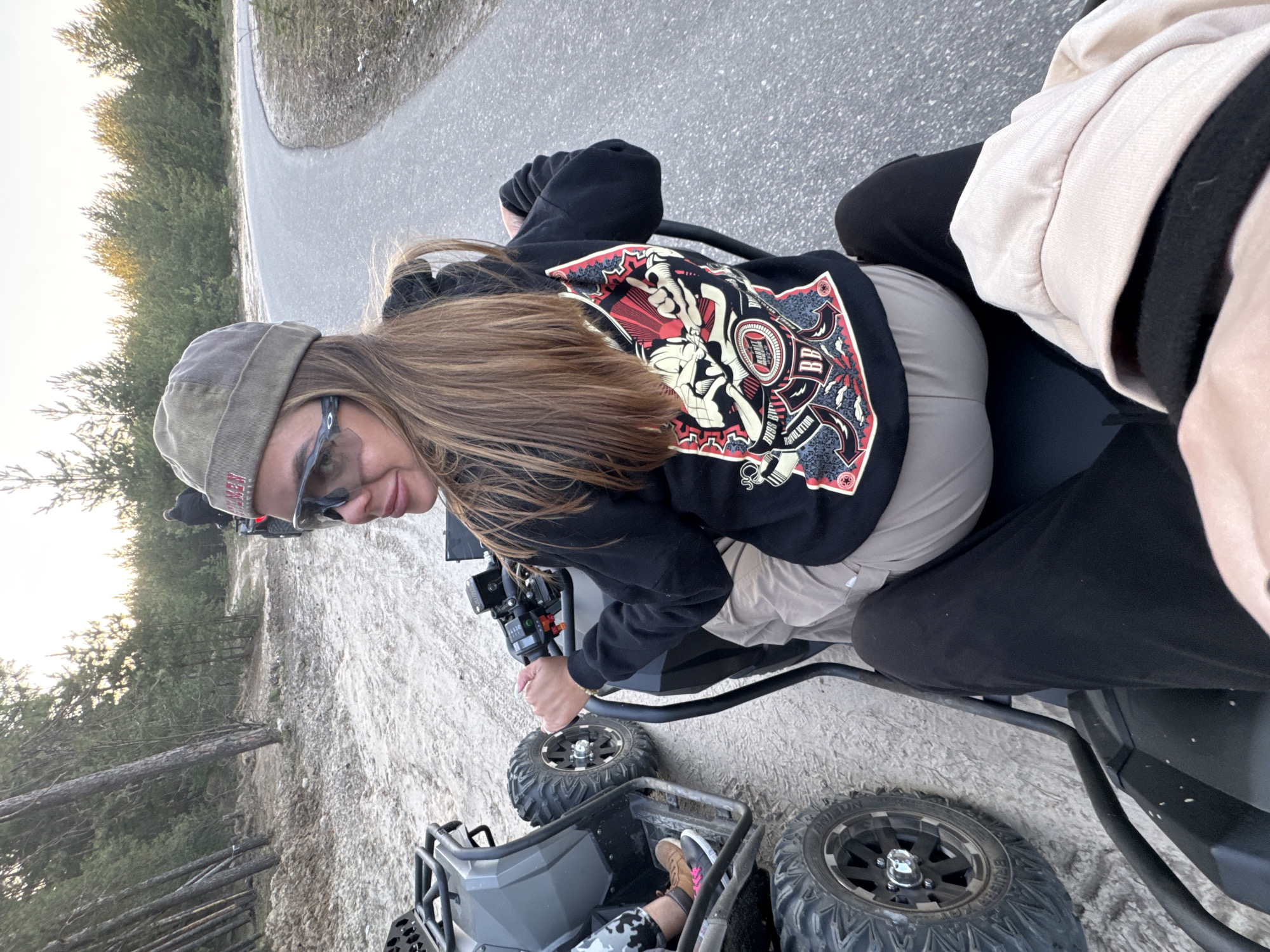 The image depicts a woman on a quad bike in Bol'shoye Kozino, Russia. She is wearing a hat, sunglasses, and a black hooded sweatshirt with a red and white graphic on the back. The quad bike is parked on a dirt surface next to a paved road. Trees are visible in the background.