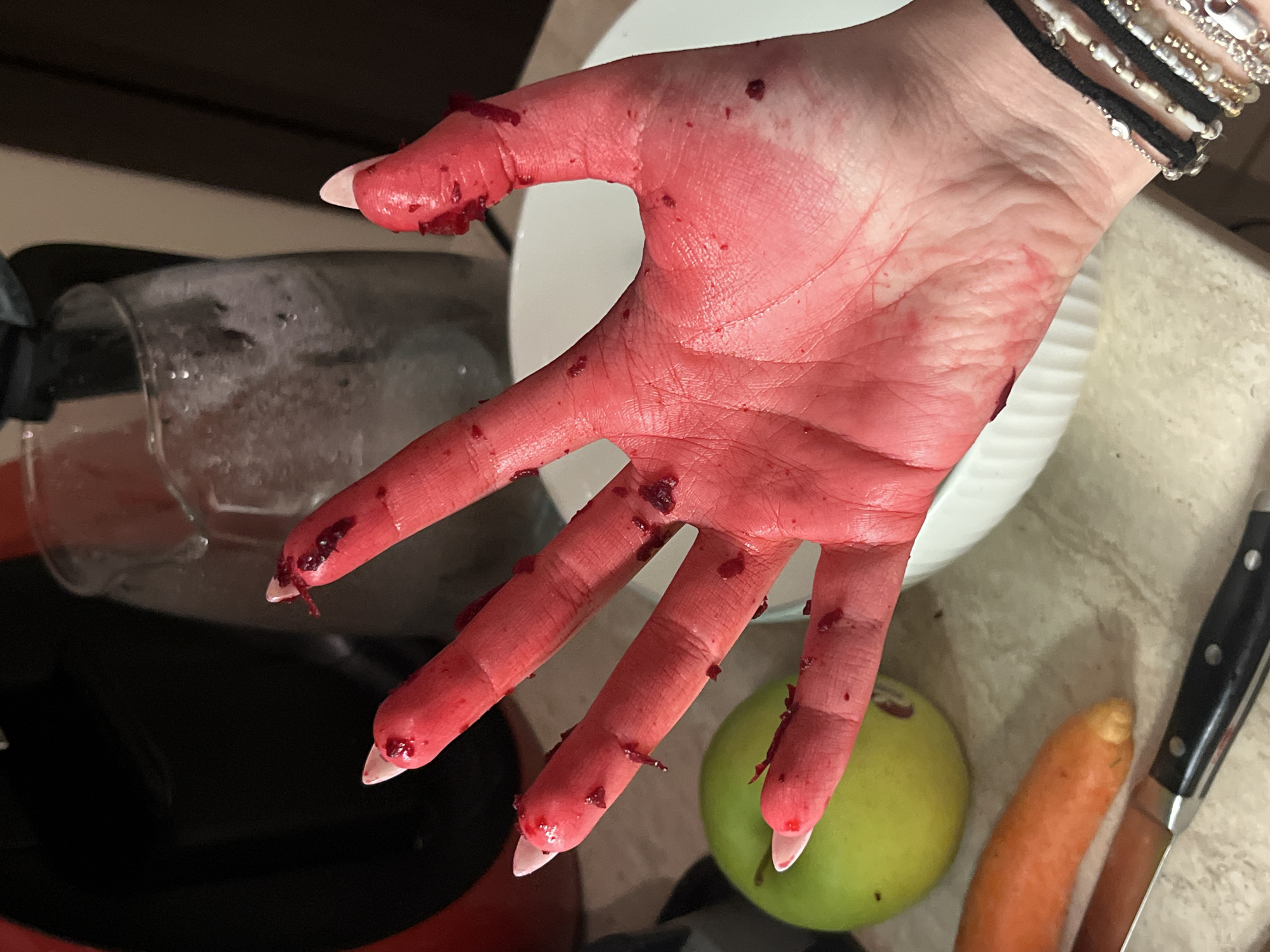 The image shows a person's hand covered in red liquid and small red pieces, possibly blood and beet pieces. The hand is in an open position, with fingers spread. The person has white manicured nails and is wearing bracelets on their wrist.
Behind the hand are various objects: a partially visible clear glass container with liquid inside, a white bowl, a green apple, an orange carrot, and a knife with a black handle. The hand, along with these objects, are positioned on what appears to be a kitchen countertop. There is no indication of the image being taken in Sofia, Bulgaria.